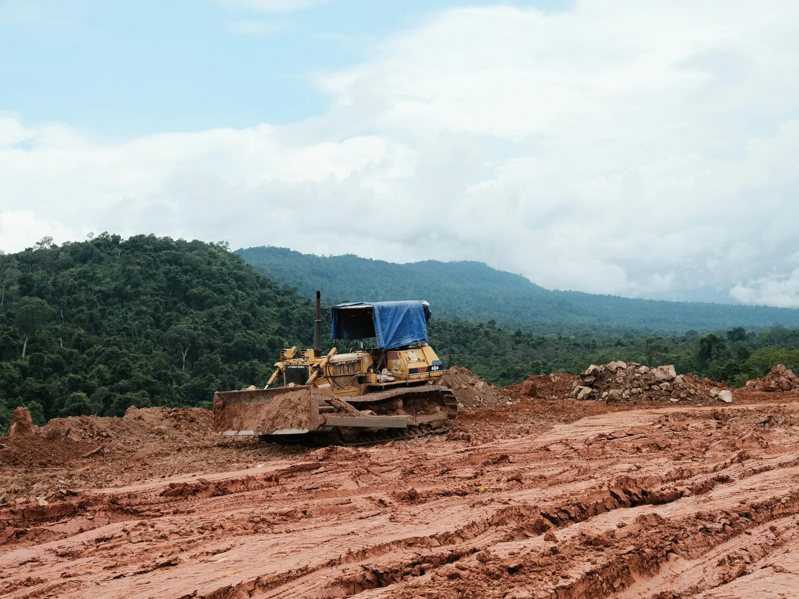 Bulldozer working on cleared red dirt land with forested hills and cloudy sky in the background.