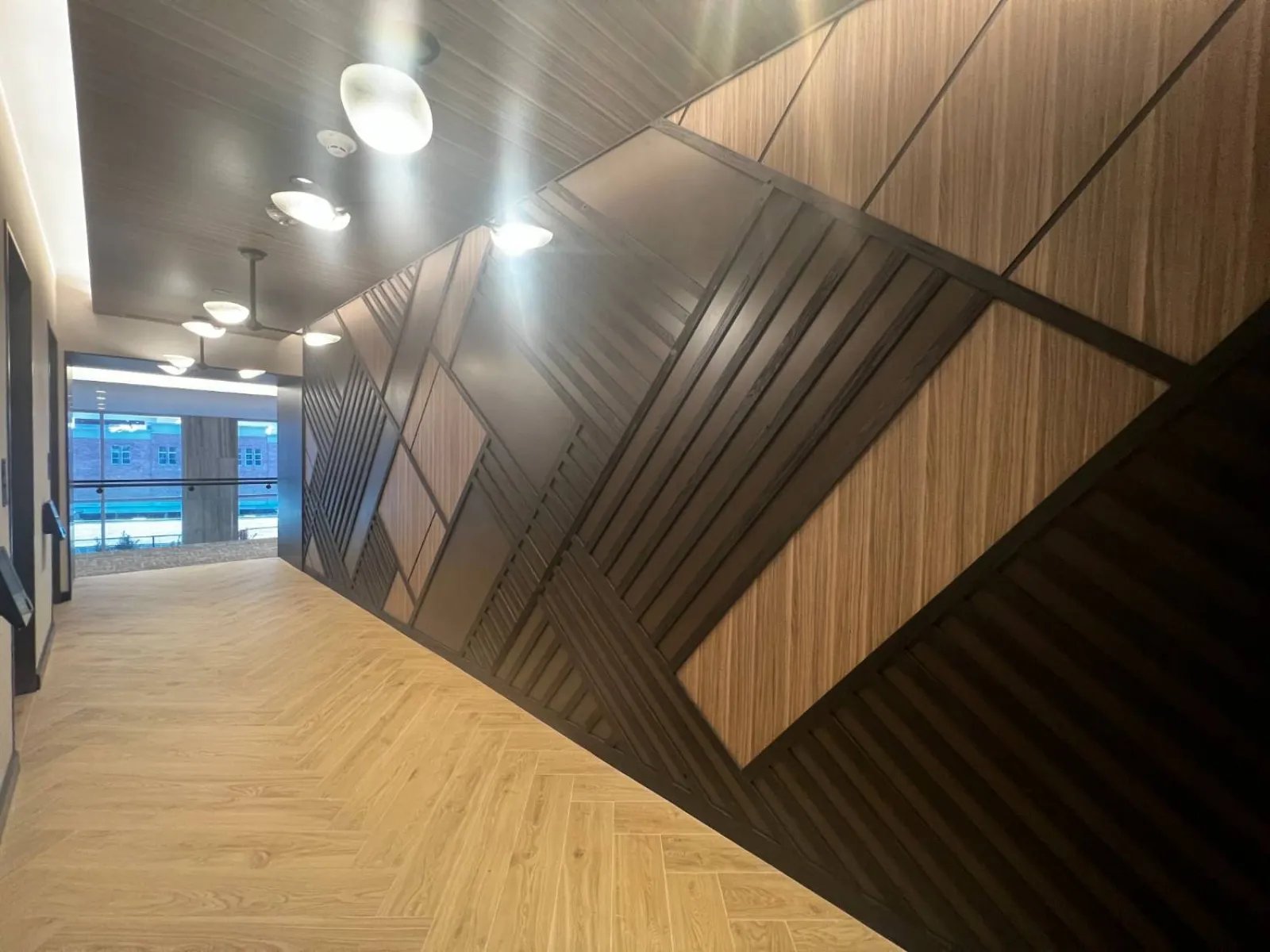 Modern hallway with geometric wood paneling, light wood flooring, and ceiling-mounted lights leading to a windowed end.