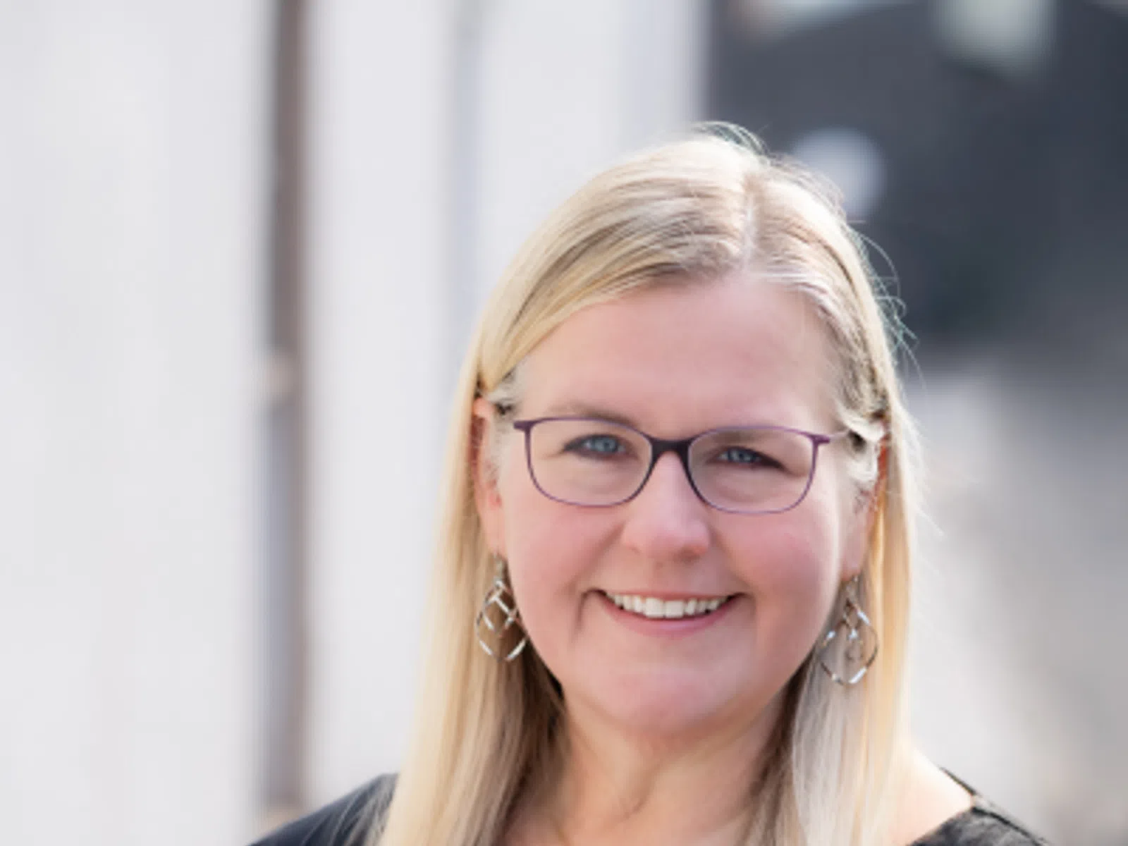 Smiling woman with long blonde hair and glasses wearing a black top and geometric earrings in natural light.