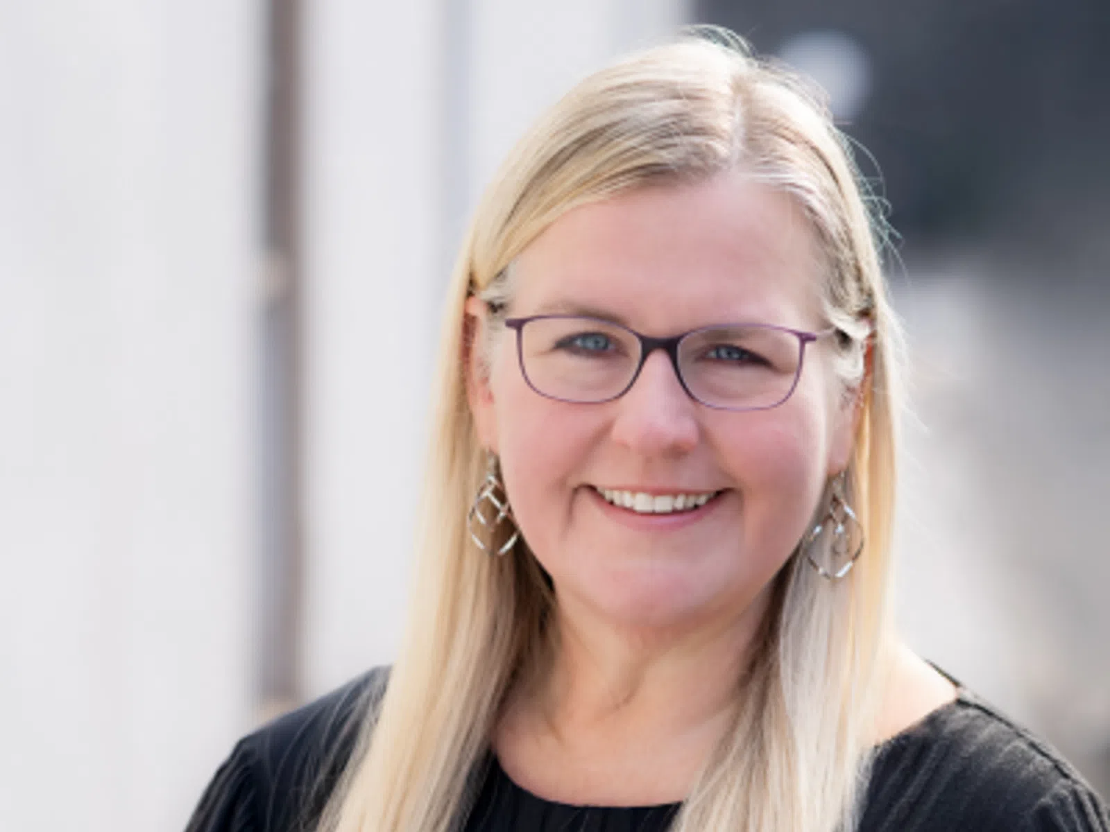 Smiling woman with long blonde hair and glasses wearing a black top and geometric earrings in natural light.