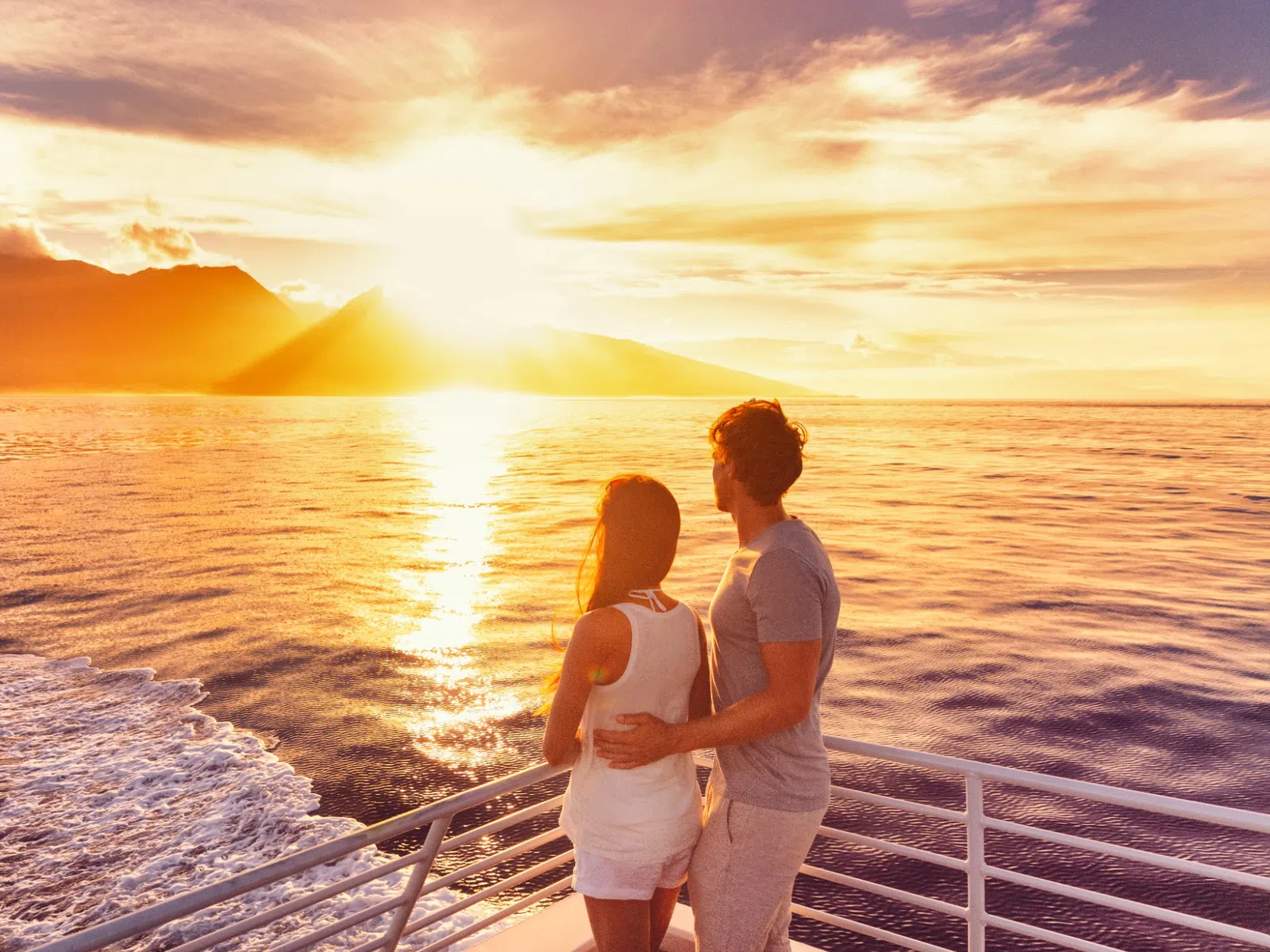 Couple on boat railing watching vibrant sunset over calm ocean and distant island mountains.