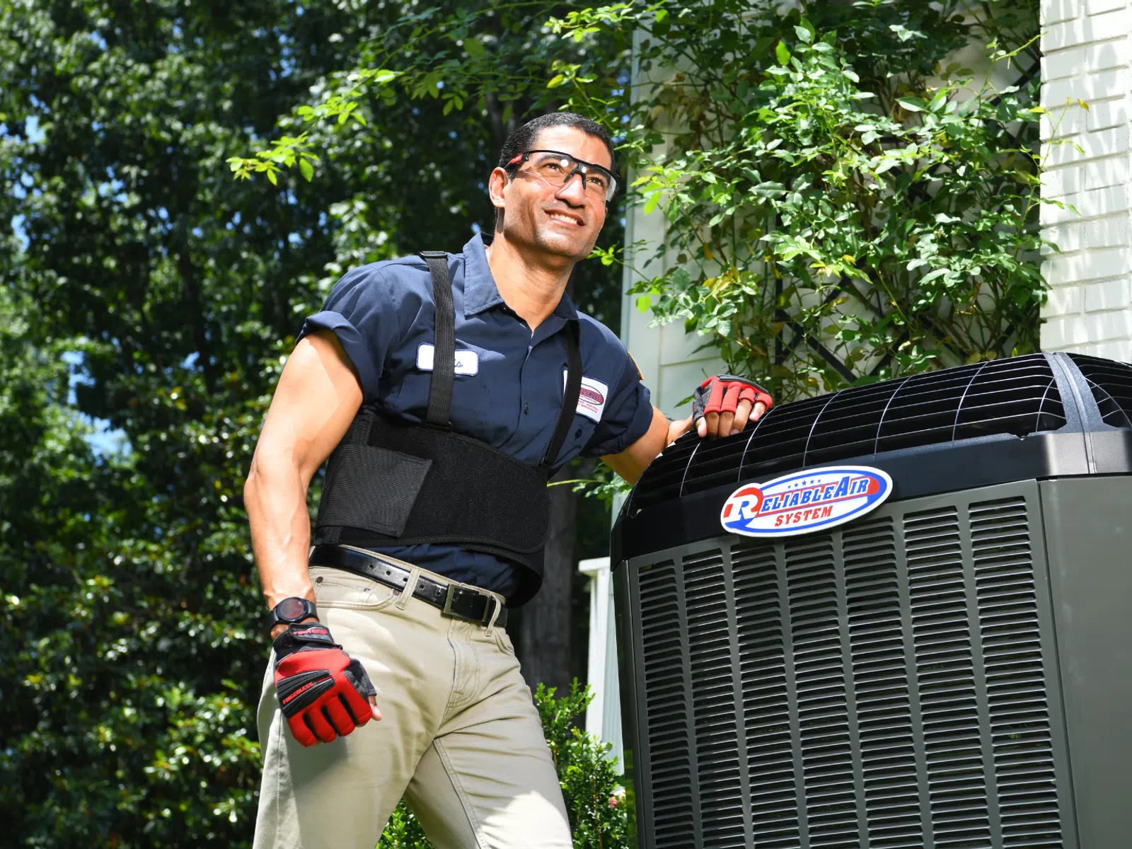 Technician wearing safety gear inspecting an outdoor air conditioning unit by a white brick house on a sunny day