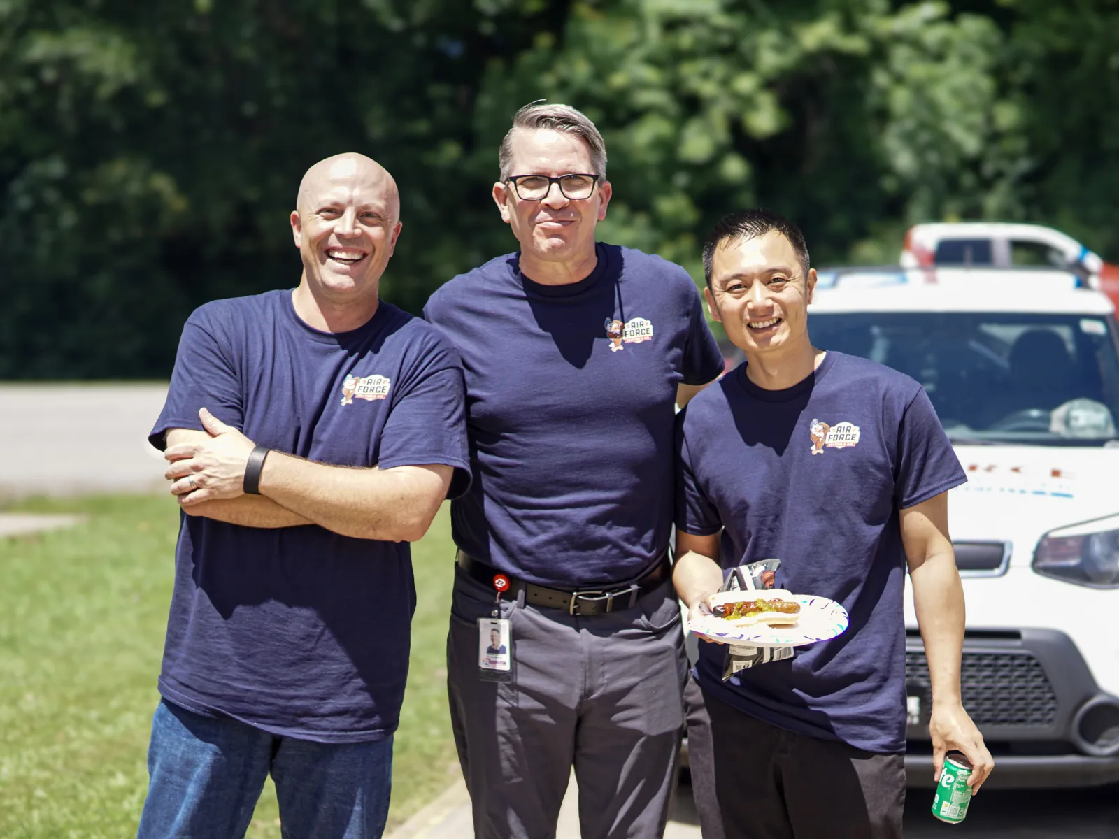 Three men in navy blue shirts pose smiling outdoors with a white vehicle in the background during a sunny day.