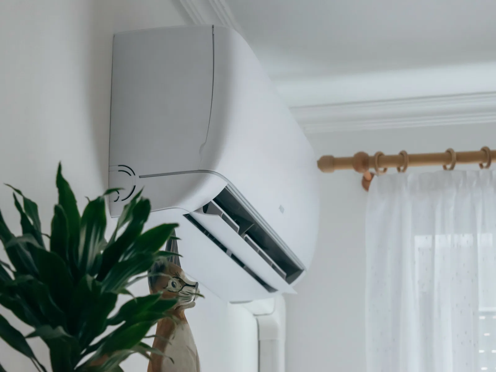 Modern white wall-mounted air conditioner installed above a window with white curtains and green plant nearby