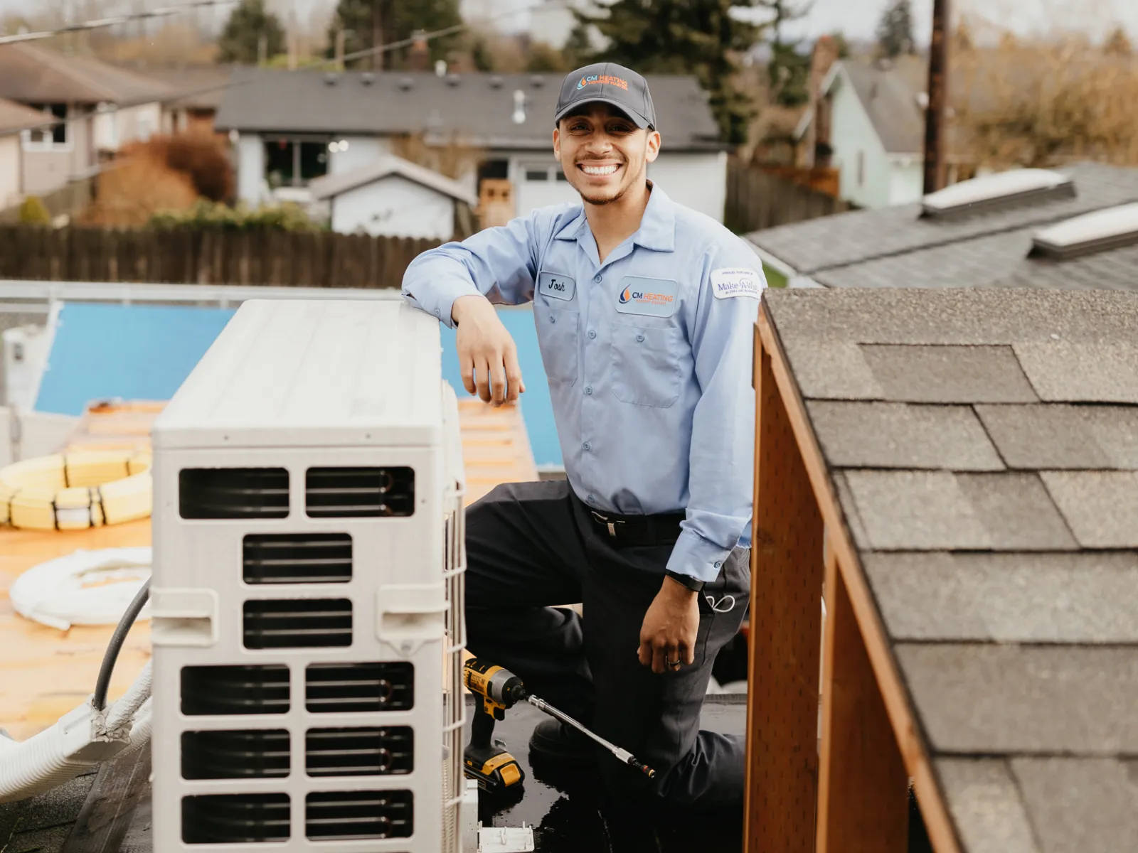 Smiling HVAC technician in uniform kneeling on a roof next to an air conditioning unit with tools in daylight.