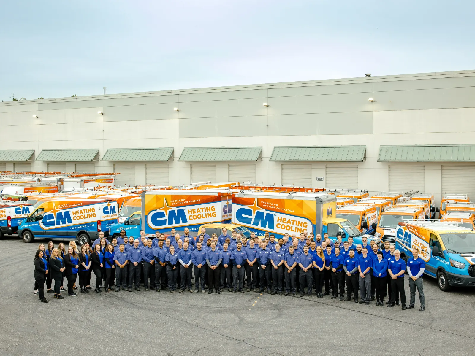 Large group of CM Heating Cooling employees posing in front of company service vans outside warehouse.
