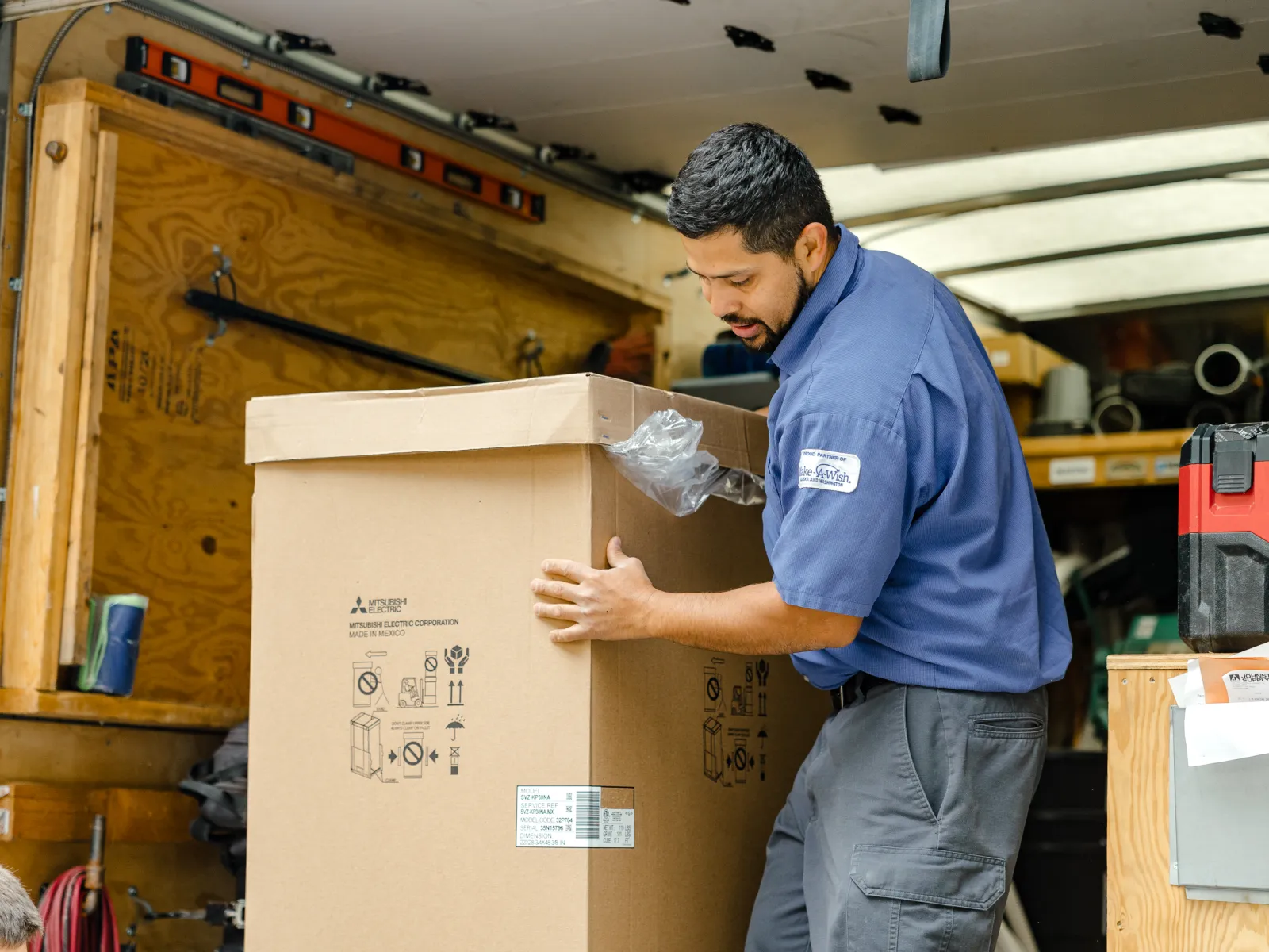 Delivery worker unloading large sealed cardboard box from a truck amid tools and equipment.