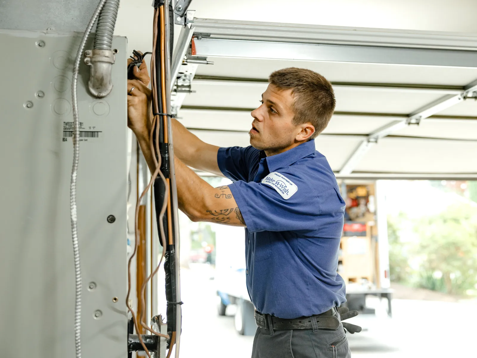 Electrician in blue uniform repairing wiring inside a residential electrical panel in a garage.