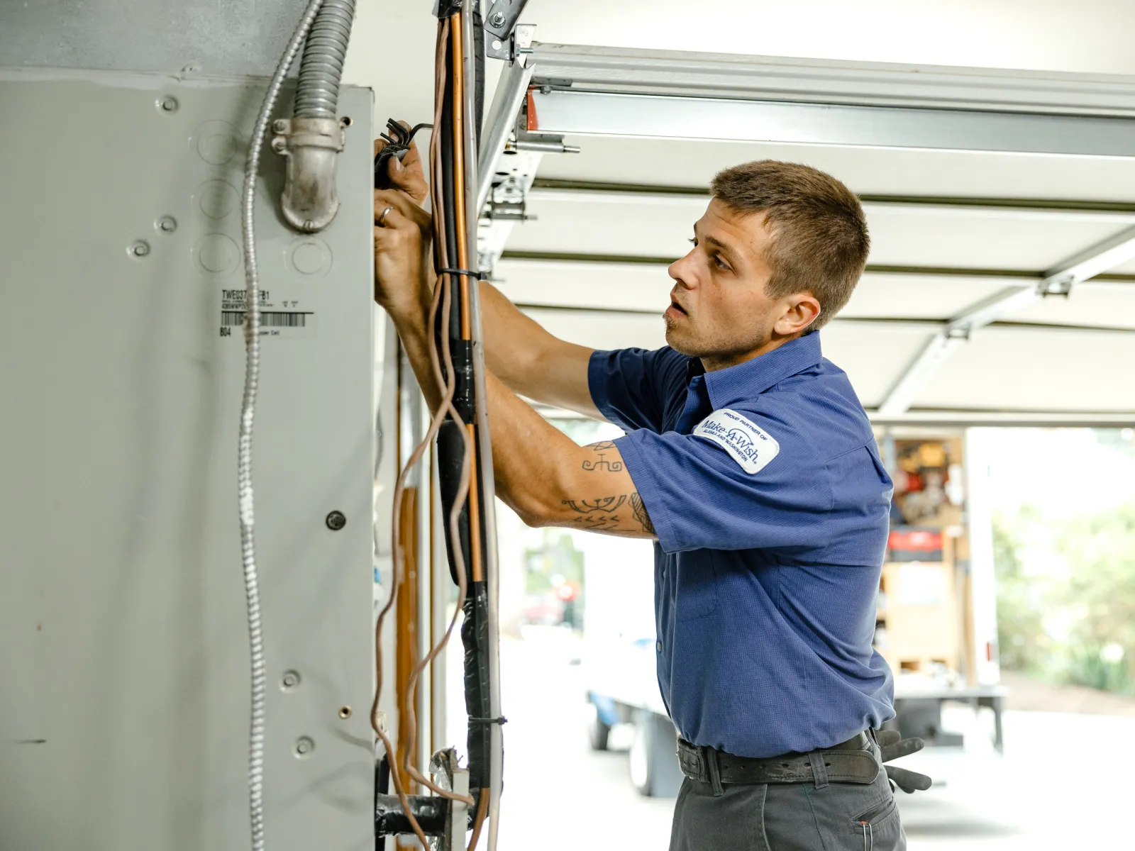 Electrician in blue uniform repairing wiring inside a residential electrical panel in a garage.
