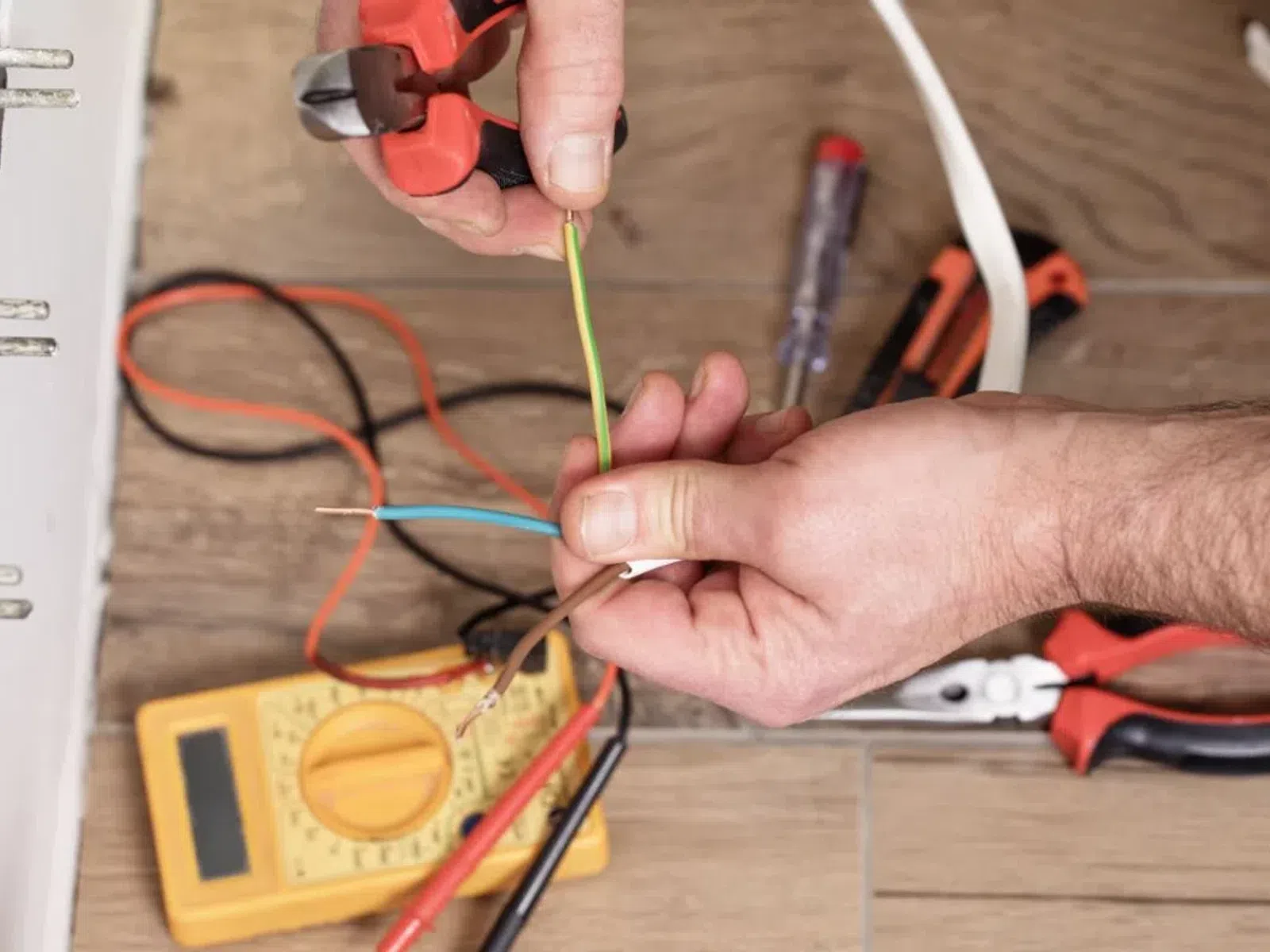 Electrician using wire cutters to strip electrical wires beside a yellow multimeter on wooden floor