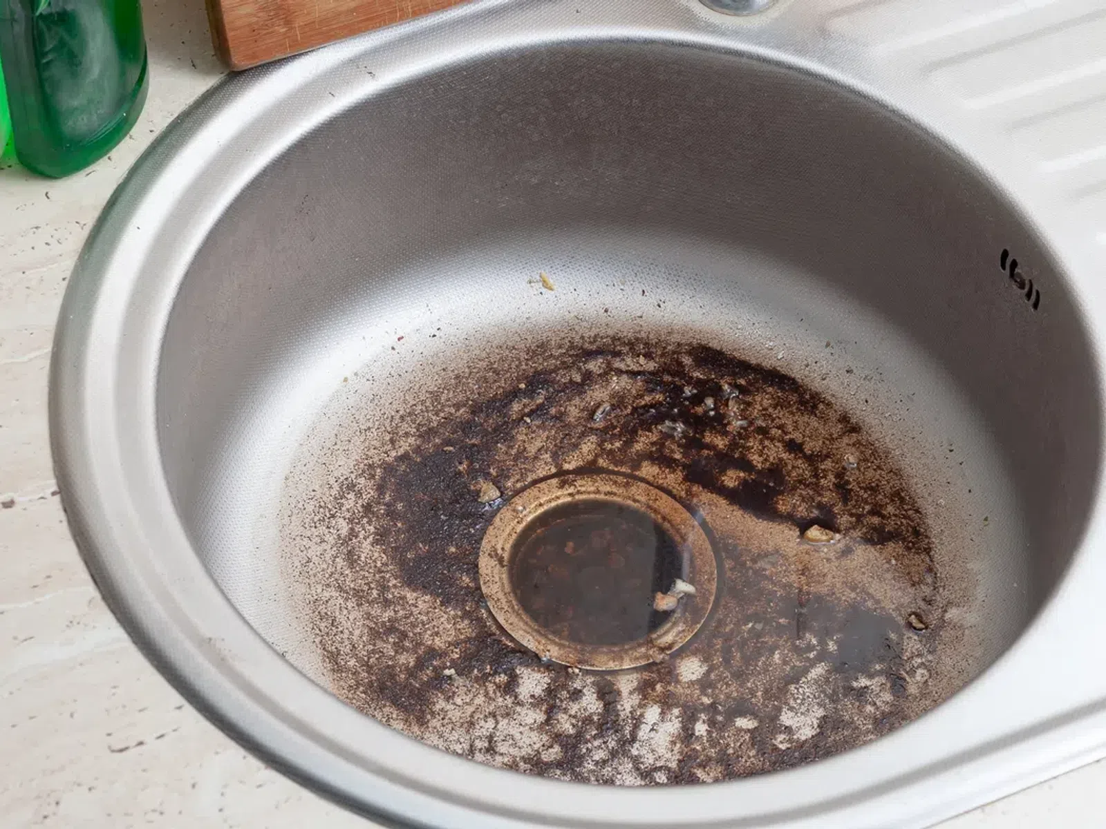 Dirty kitchen sink with food residue and dark stains around the drain area awaiting cleaning.
