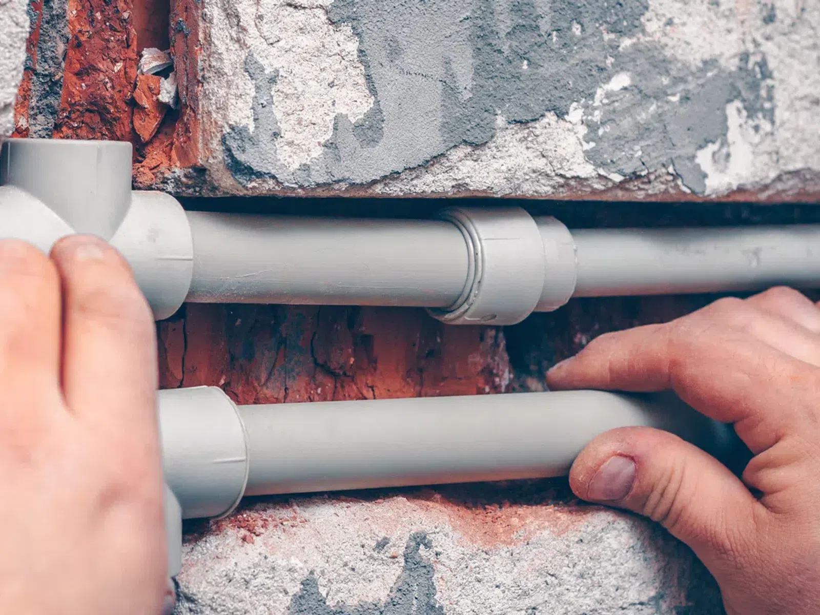 Hands installing gray PVC pipes on a brick wall during plumbing work or construction project.