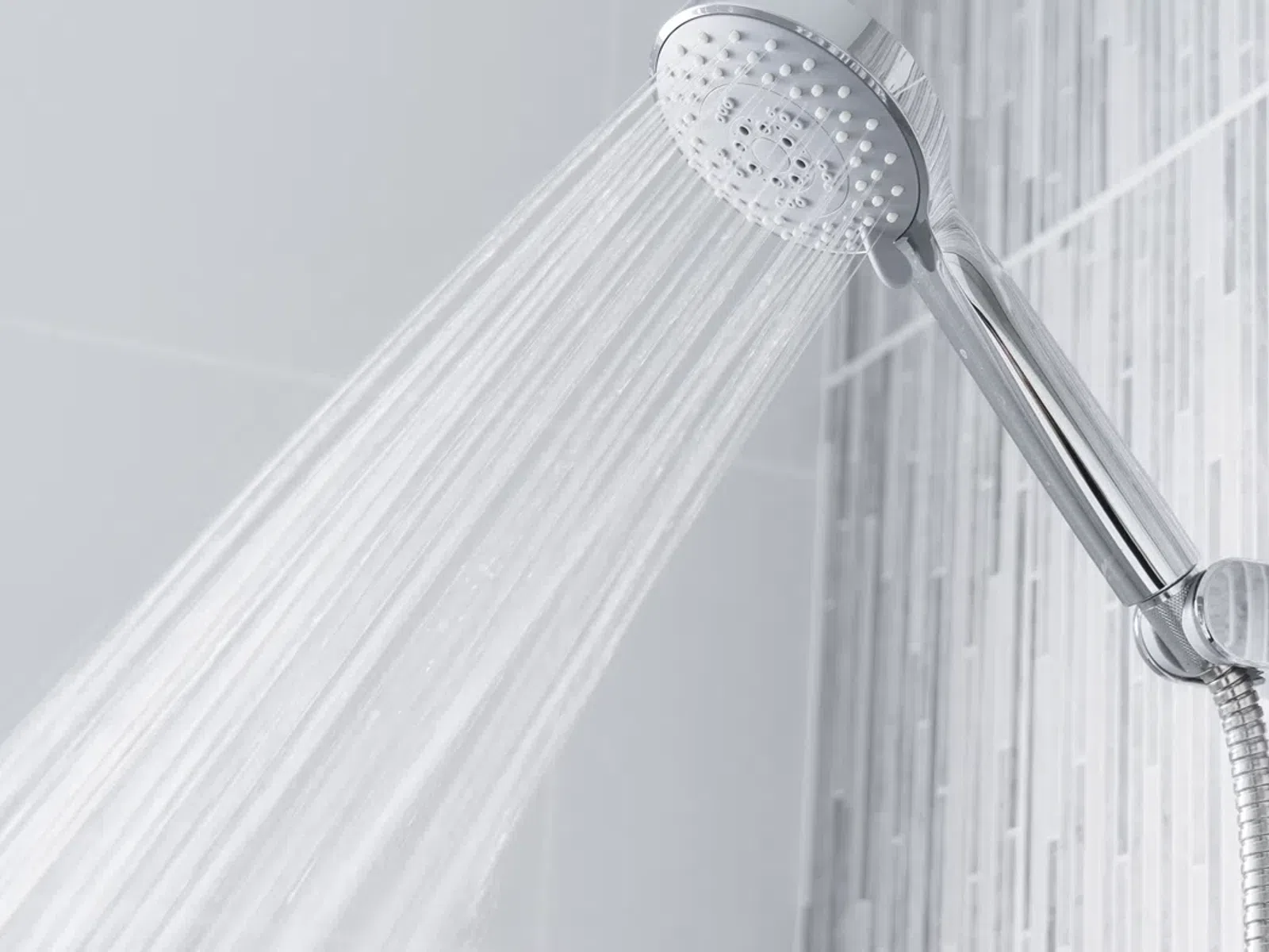 Chrome handheld showerhead spraying water in a modern tiled bathroom with vertical gray tiles.