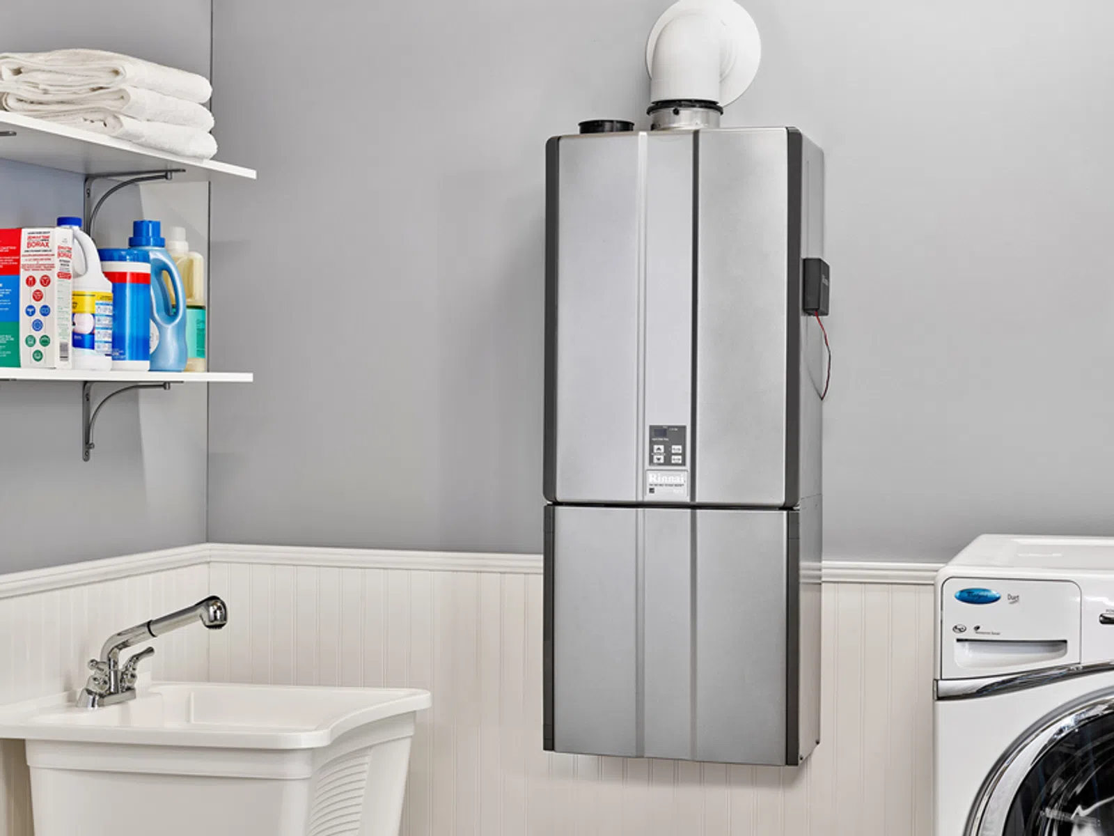Modern laundry room with wall-mounted silver water heater, washer, sink, and cleaning supplies on shelves.