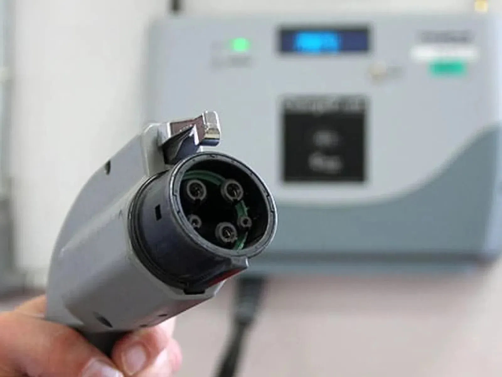 Hand holding an electric vehicle charging plug with a charging station blurred in the background.