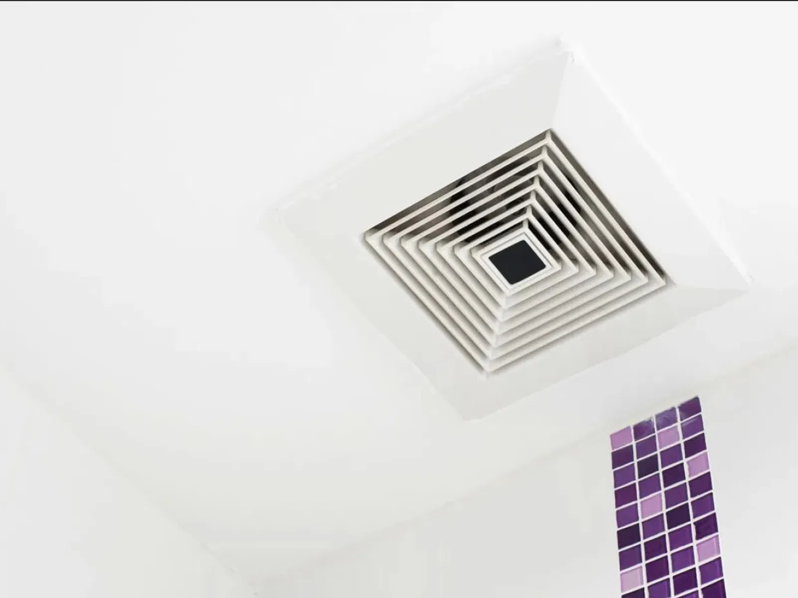 Ceiling air vent with white louvered grille and purple mosaic tile strip on white wall.
