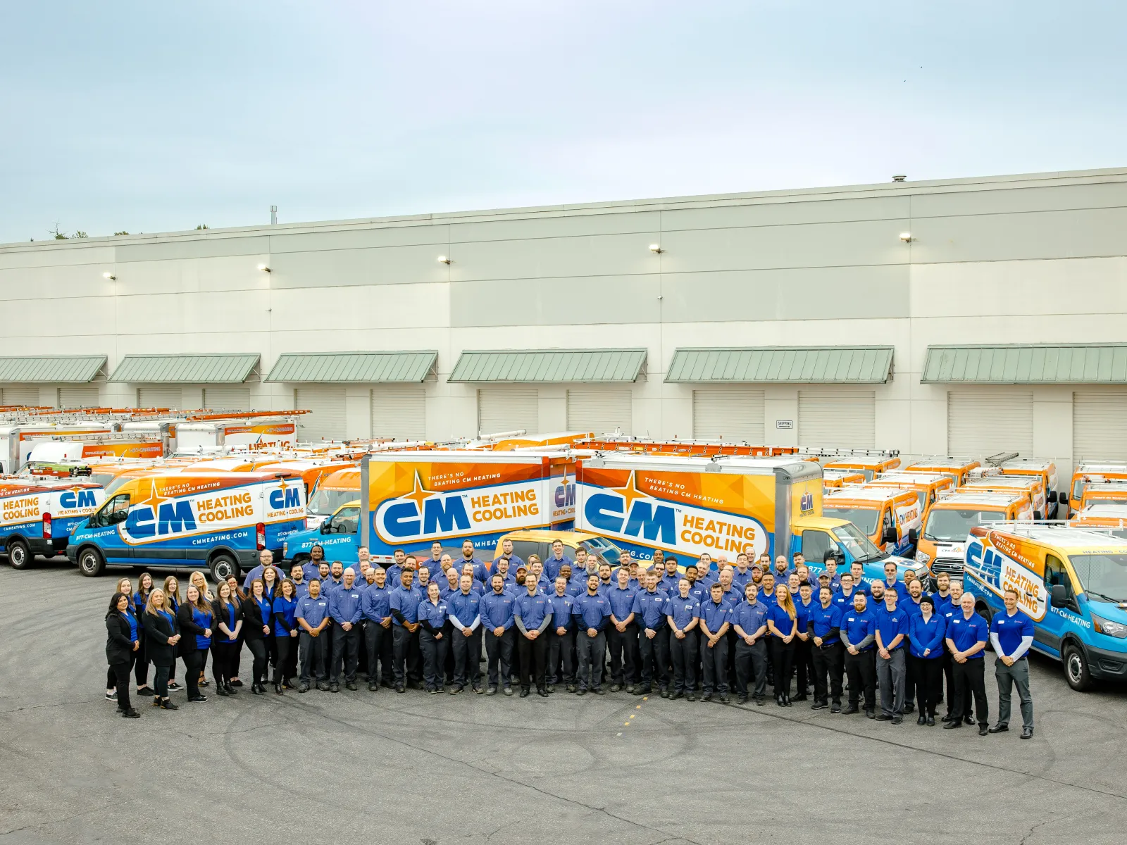 Large team of employees standing in front of a fleet of CM Heating and Cooling service trucks outside a warehouse.