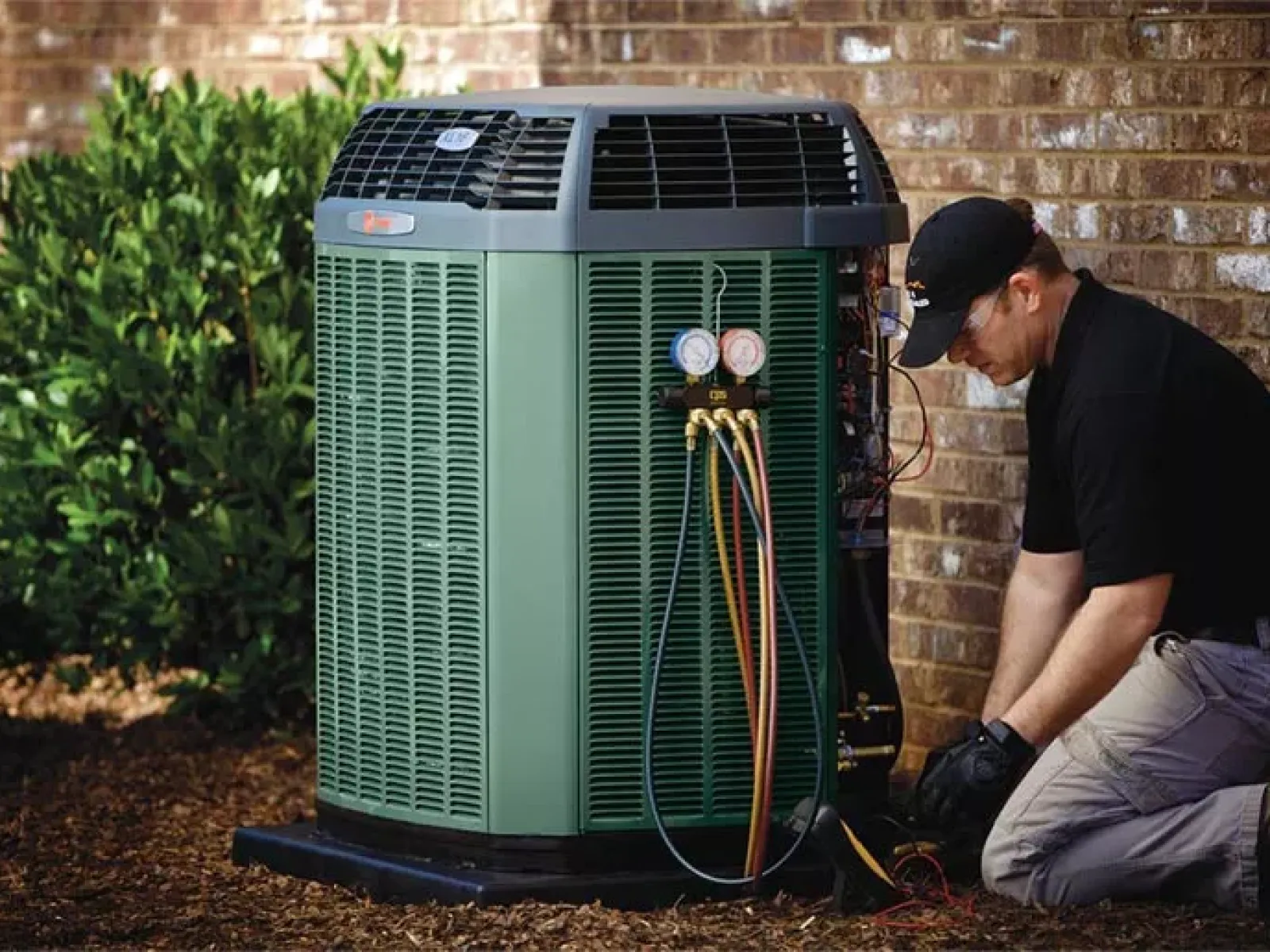 Technician repairing a green outdoor air conditioning unit next to a brick wall with diagnostic gauges attached