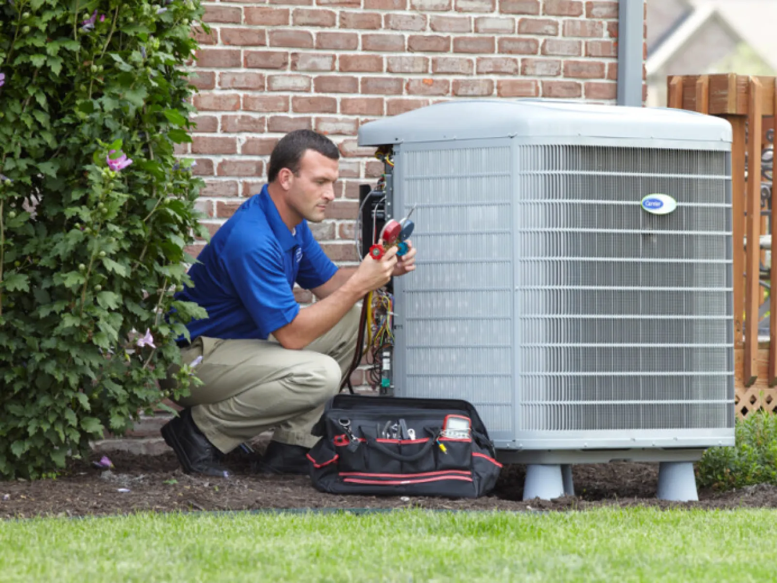 Technician repairing an outdoor air conditioning unit beside a brick wall with a tool bag on the ground.