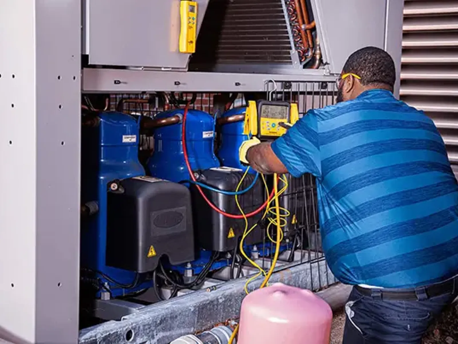 Technician in blue striped shirt testing HVAC compressor unit using diagnostic equipment outdoors.