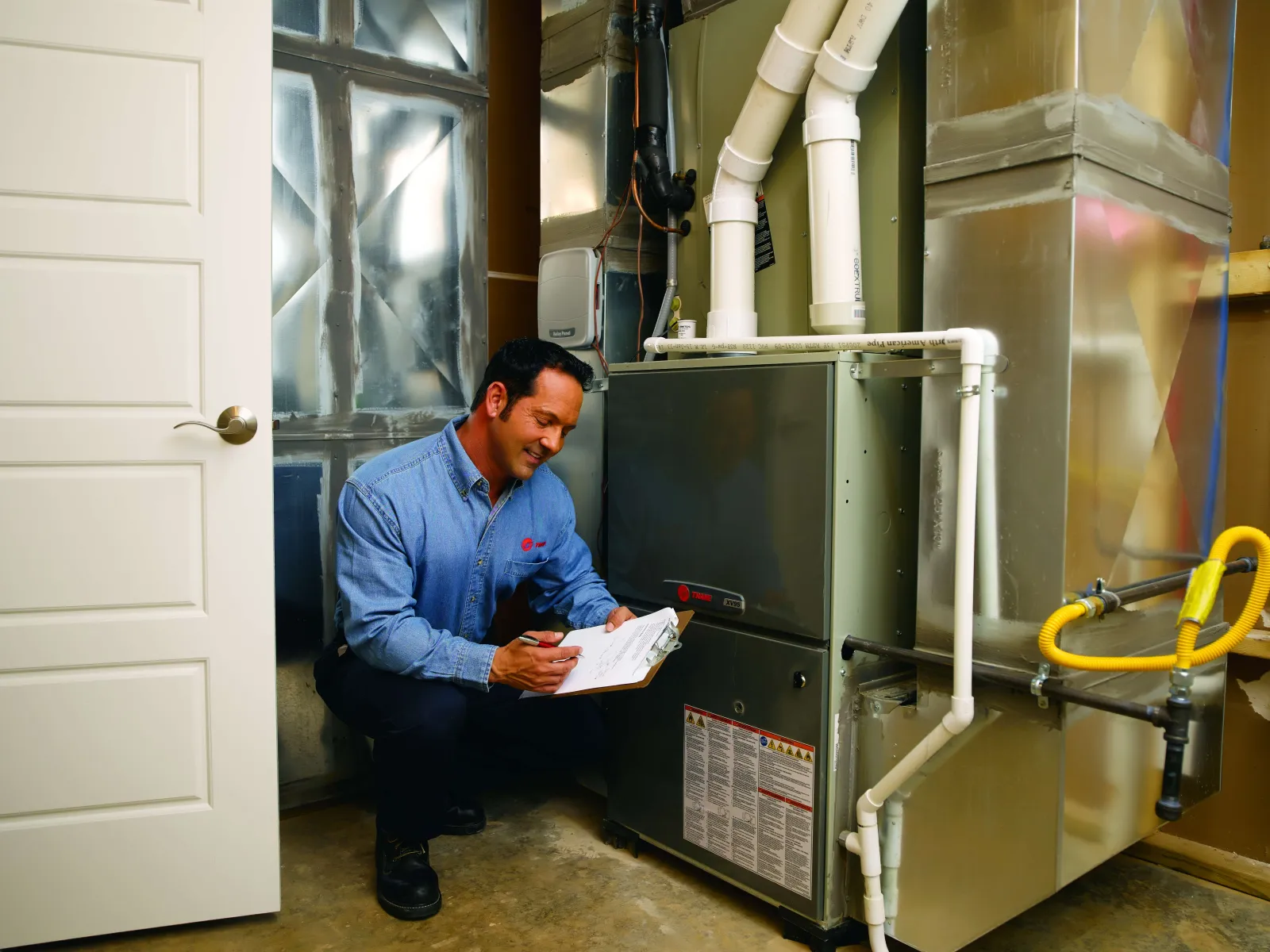 Technician inspecting and taking notes on a residential furnace system in a basement utility room.
