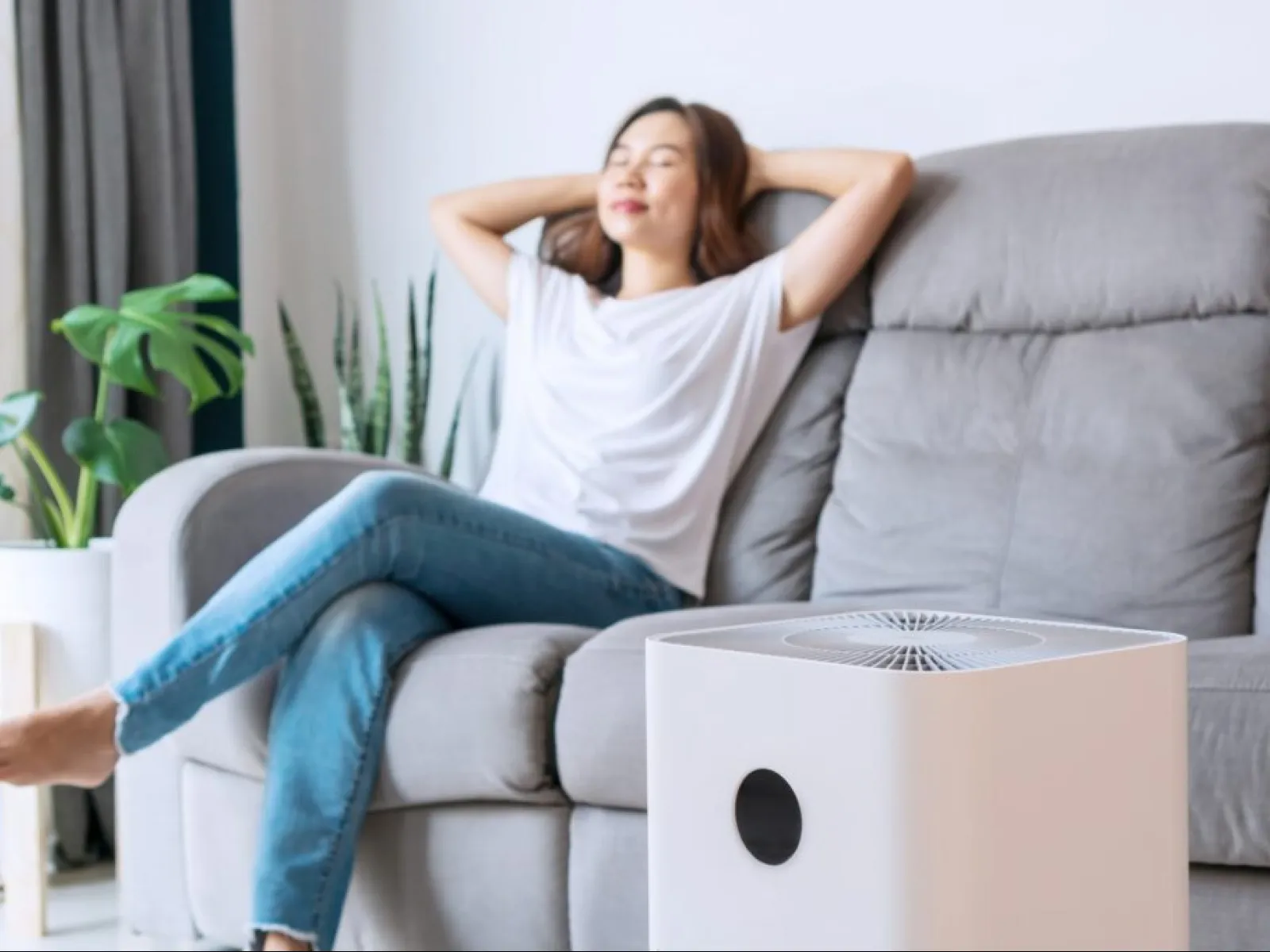 Woman relaxing on a gray sofa with a modern white air purifier in a bright living room with green plants.