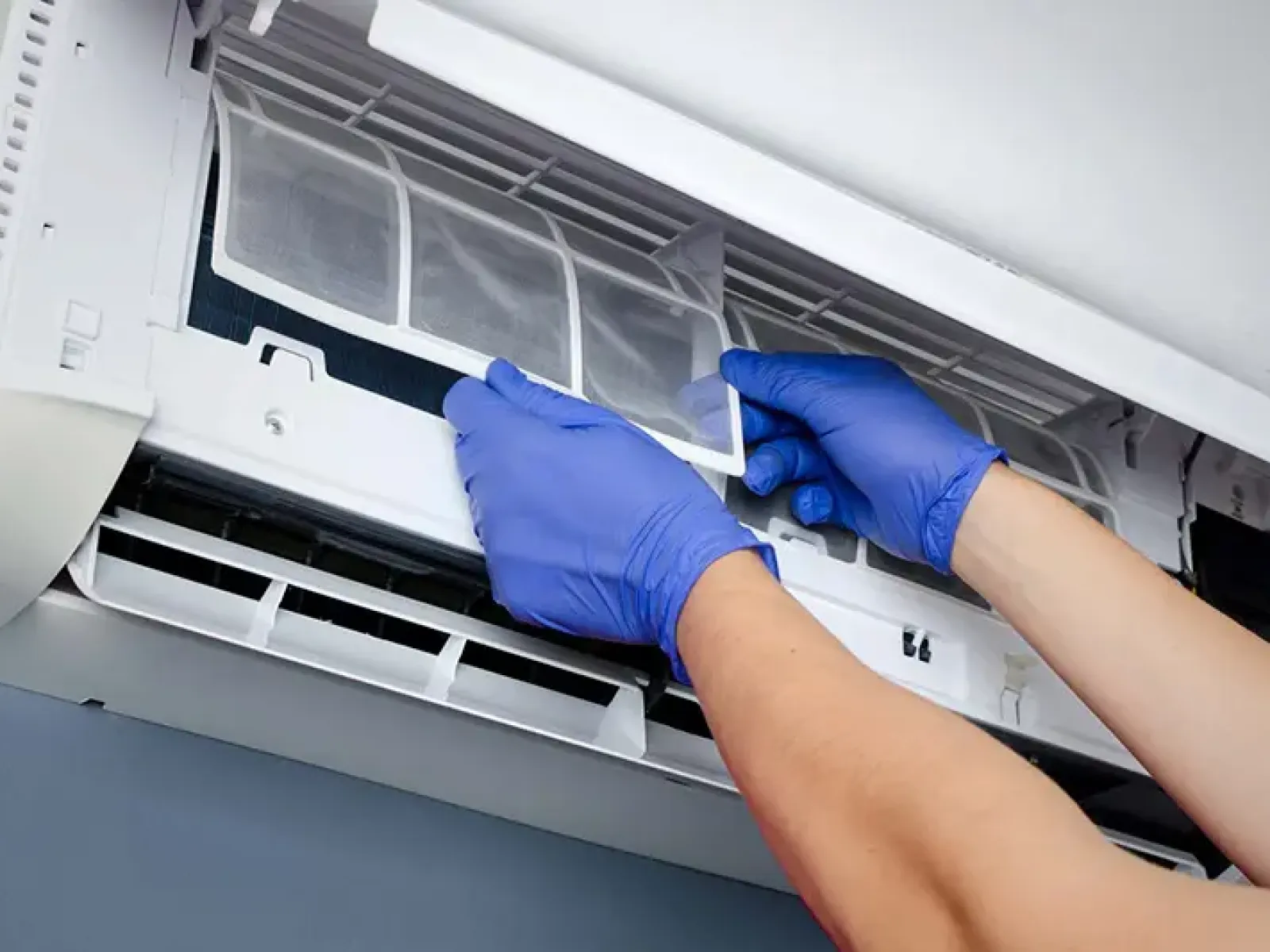 Hands with blue gloves removing or cleaning air conditioner filter in a white wall-mounted unit.