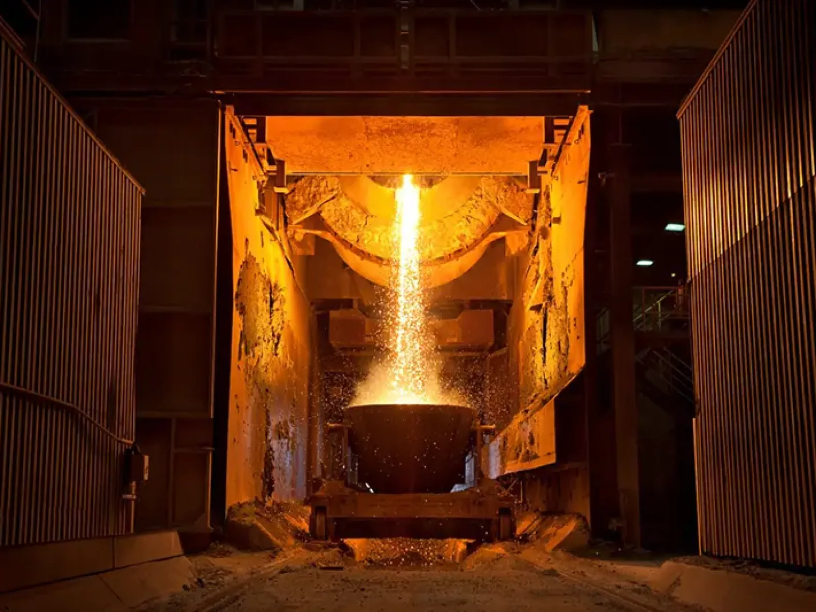 Molten metal being poured from a furnace into a crucible inside an industrial foundry with orange lighting.