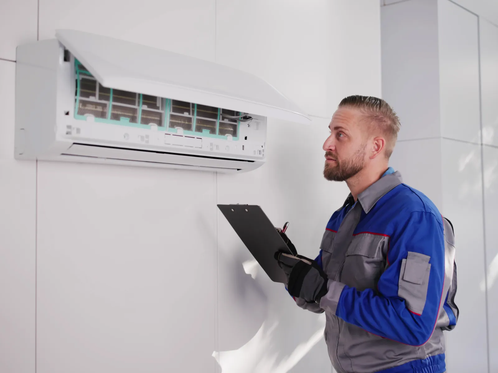 Technician in blue uniform inspecting and repairing a wall-mounted air conditioning unit with a clipboard in hand.