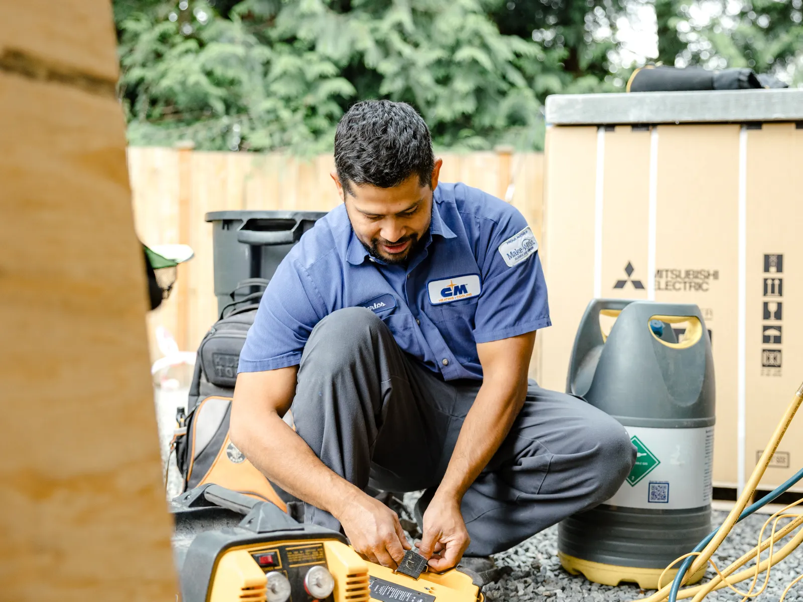 Technician in blue uniform crouching and checking equipment outdoors near HVAC gas cylinders and machinery.