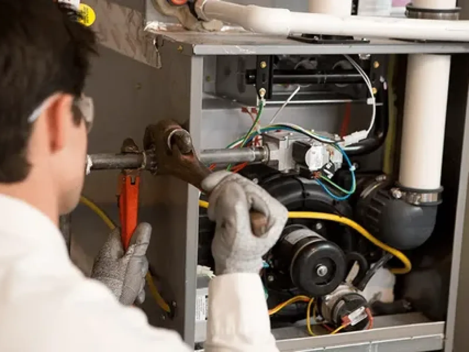 Technician wearing gloves and safety glasses repairing a furnace with pipe wrench and pliers indoors.