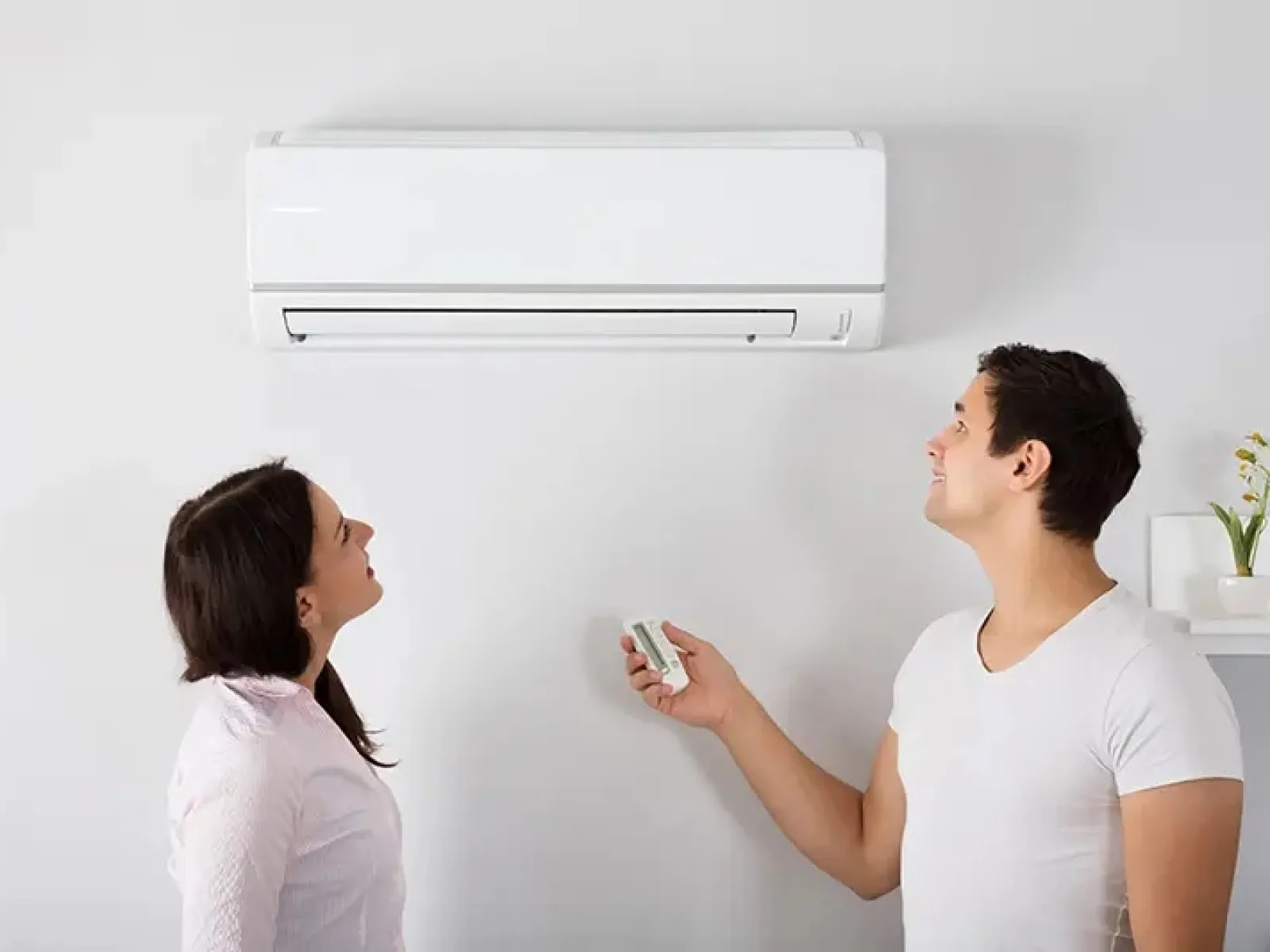 Man and woman controlling a wall-mounted air conditioner with a remote in a bright room.