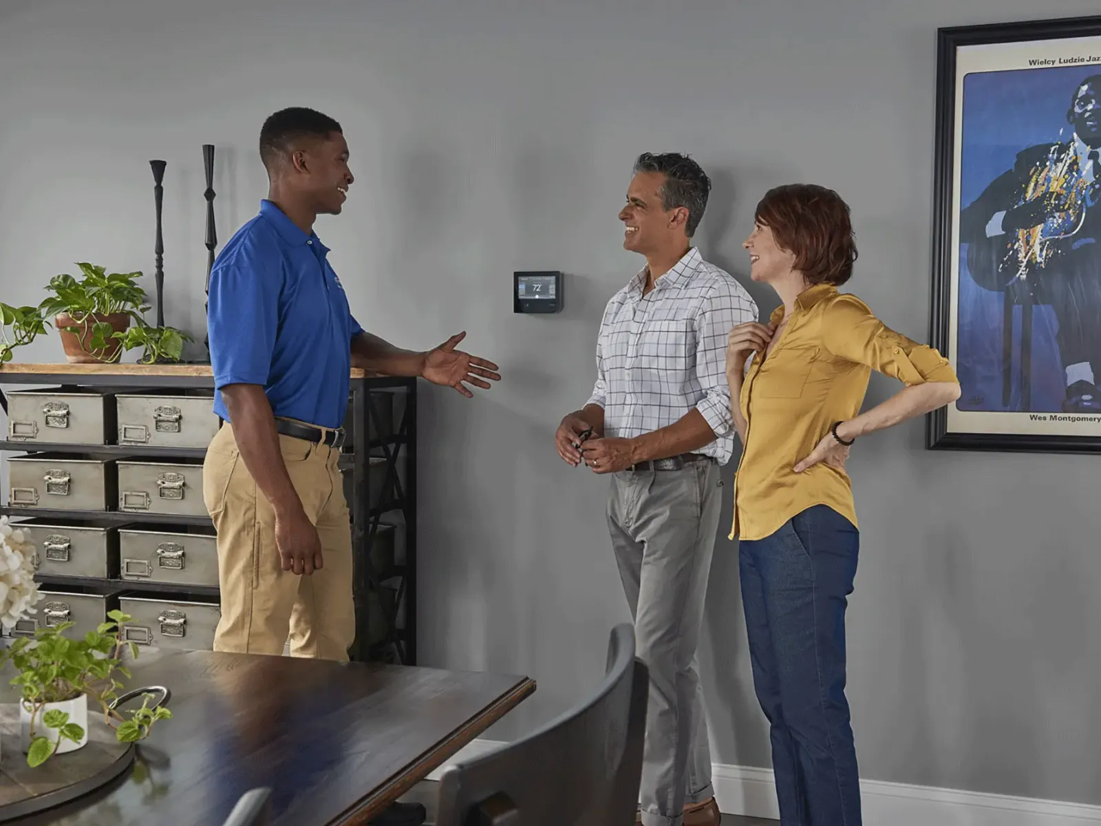 Technician in blue shirt greets couple in home with gray walls and jazz poster, discussing smart thermostat on wall.
