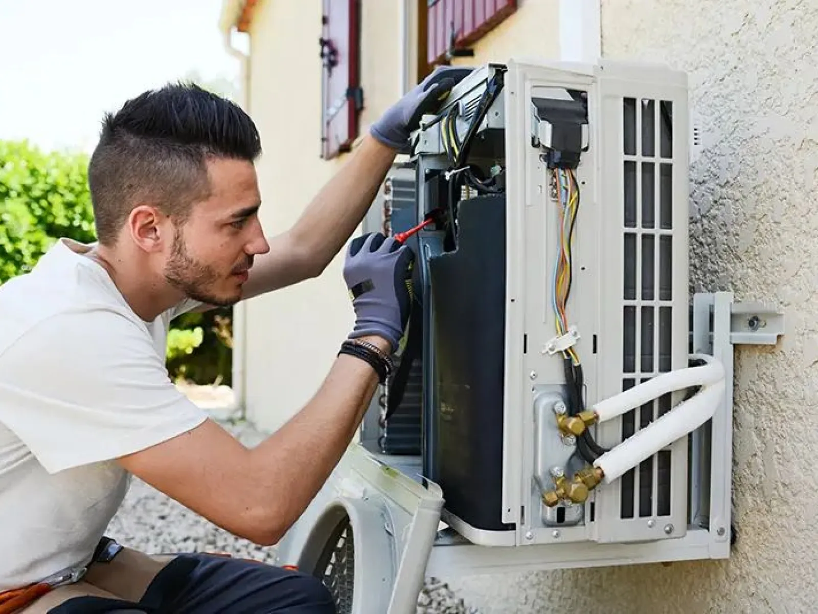 Technician repairing outdoor air conditioning unit mounted on wall using tools and wearing gloves.