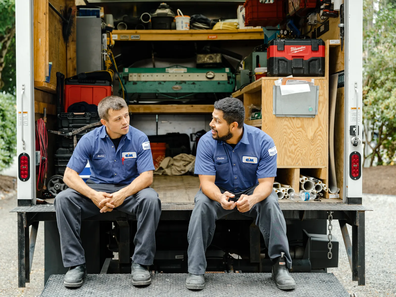 Two technicians in blue uniforms sitting at the back of a work truck having a conversation during a break.