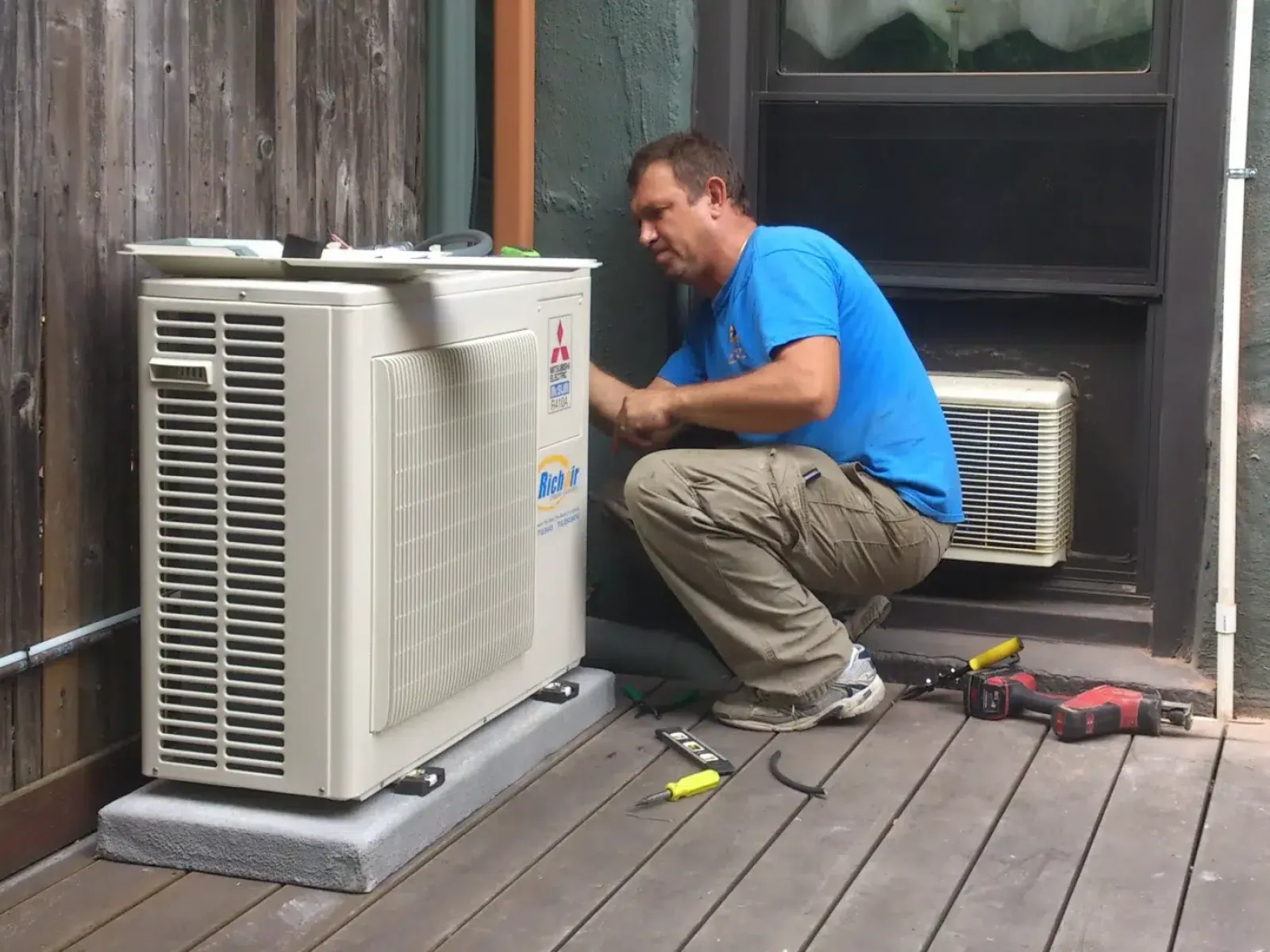 Technician installing or repairing an outdoor air conditioning unit on a wooden deck beside a house.
