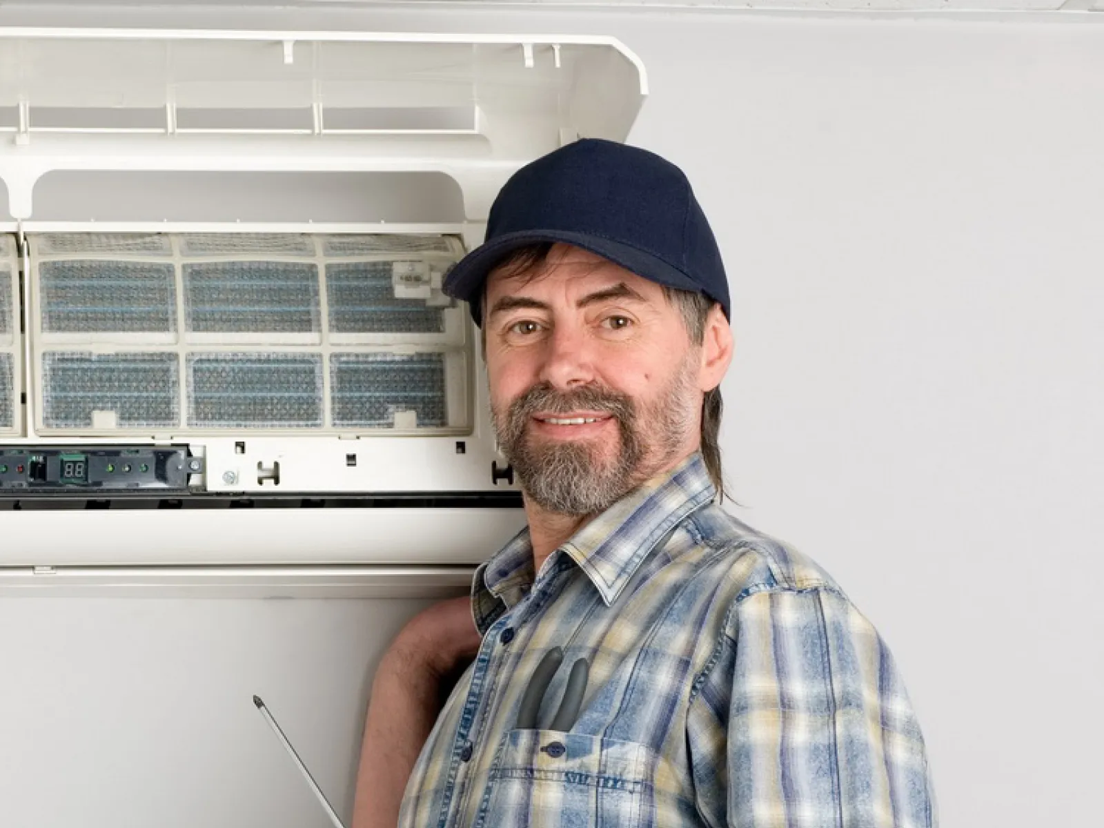 Smiling technician in plaid shirt and cap repairing a wall-mounted air conditioner with a screwdriver.