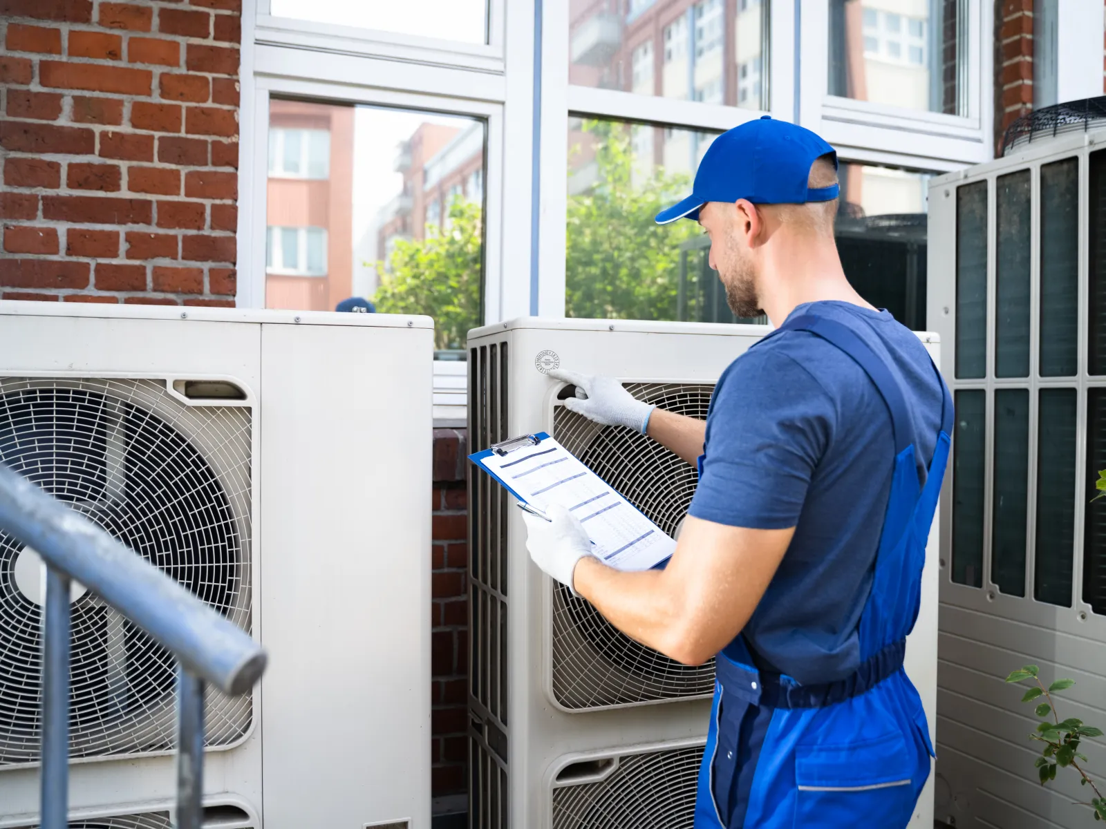 Technician in blue uniform inspecting outdoor air conditioning units with clipboard near brick wall and windows.