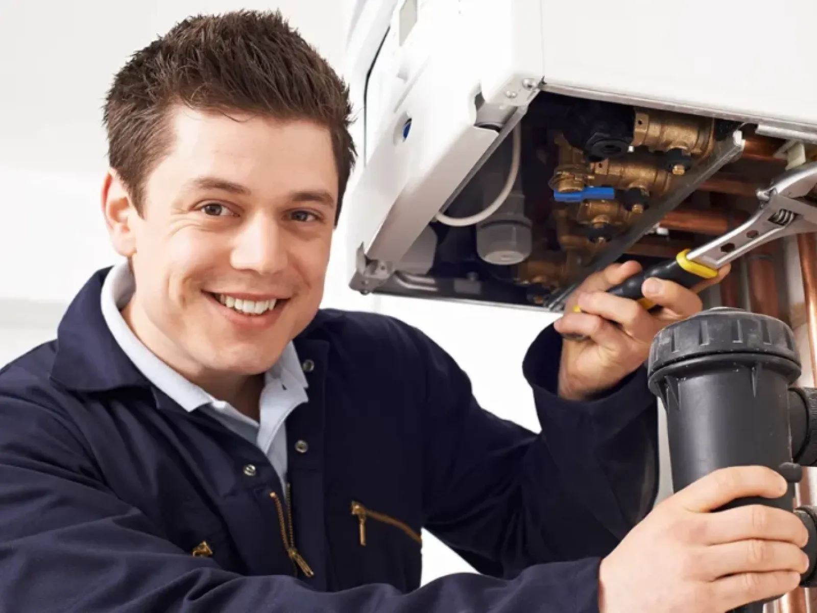 Smiling plumber in navy coveralls fixing boiler pipes with wrench in indoor utility room
