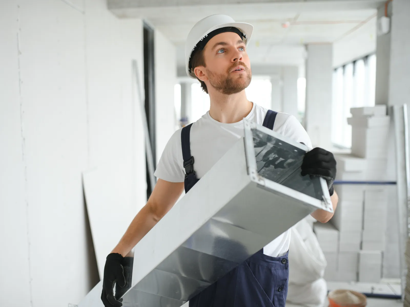 Construction worker wearing a helmet carrying a large metal duct inside a building under construction.