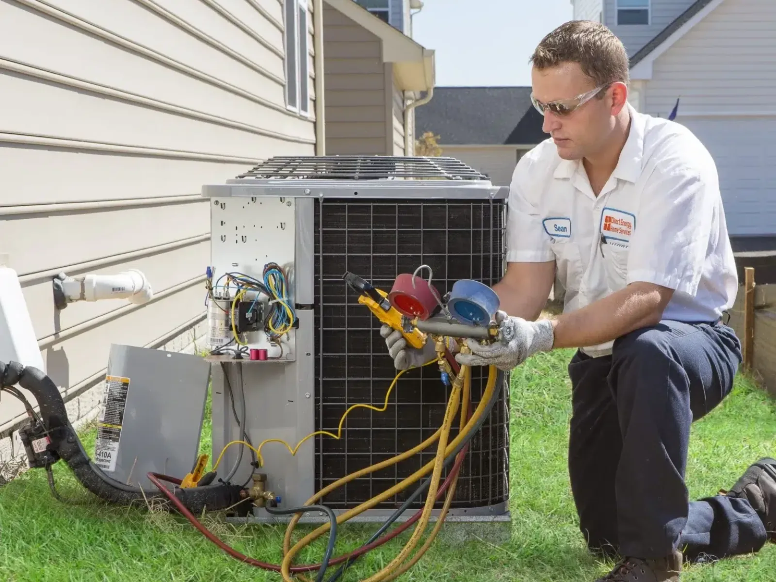 Technician in uniform inspecting and repairing an outdoor air conditioning unit on a lawn beside a house.