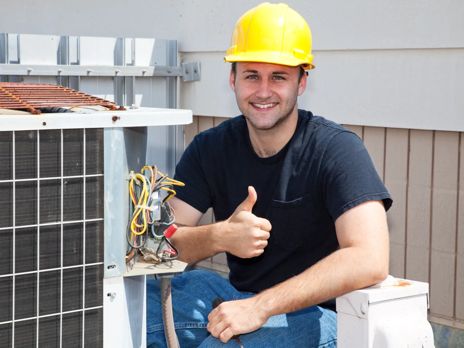 HVAC technician in yellow hard hat giving thumbs up while working on outdoor air conditioning unit.
