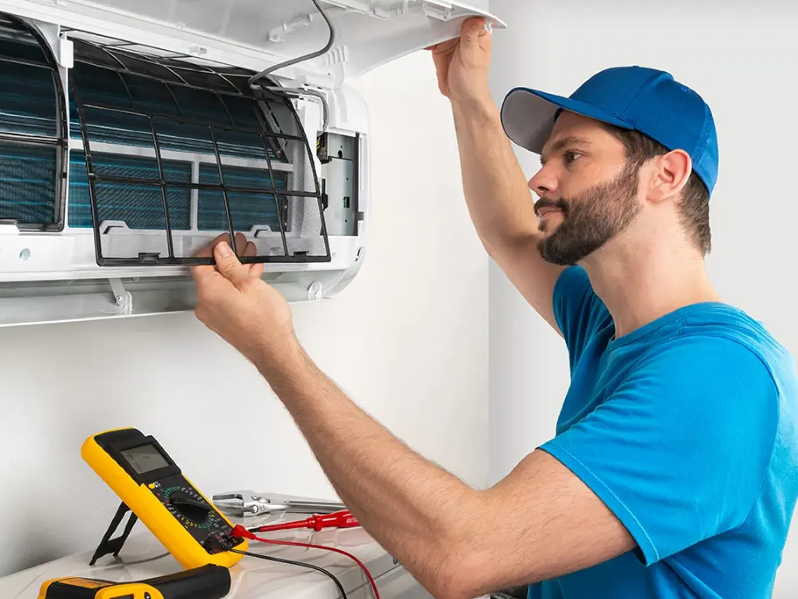 Technician in blue shirt and cap repairing air conditioner unit with multimeter and tools nearby indoors.