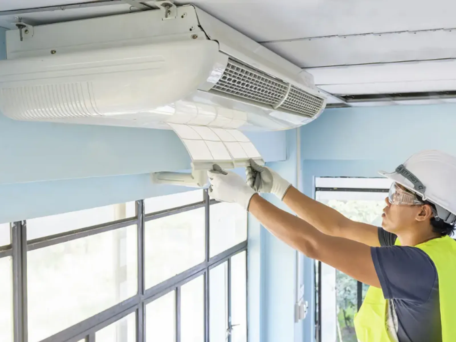 Technician wearing safety gear installs or repairs a white ceiling-mounted air conditioning unit in a bright room.