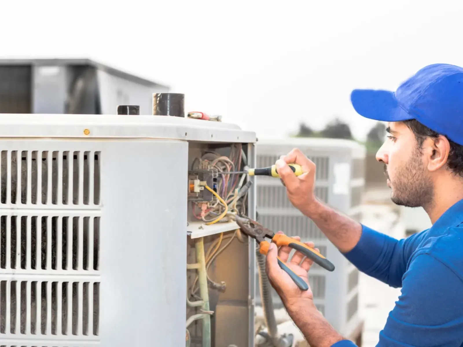 Technician in blue uniform repairing outdoor air conditioning unit using pliers and screwdriver