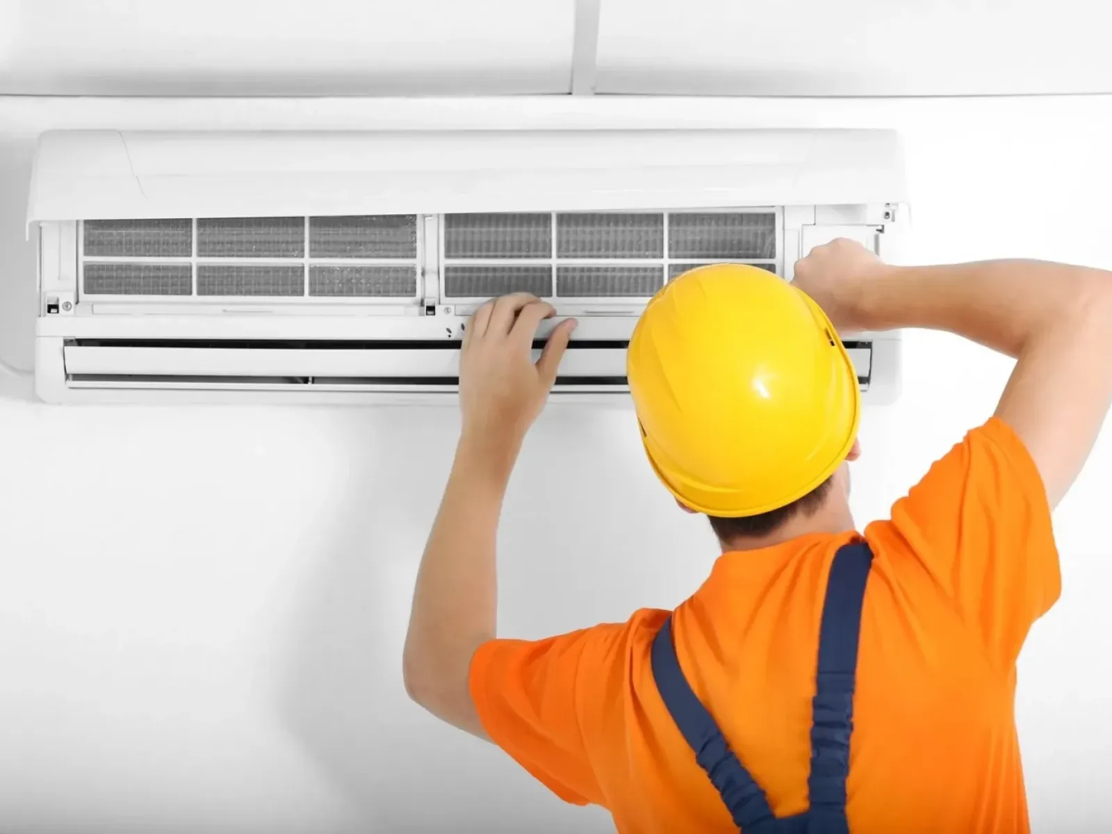 Technician in yellow helmet and orange shirt repairing or installing a wall-mounted air conditioner indoors.