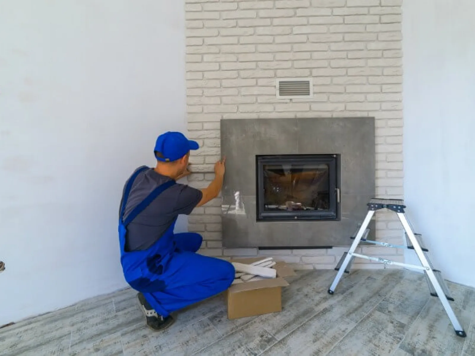 Worker in blue overalls installing or inspecting fireplace in a modern living room with white walls.
