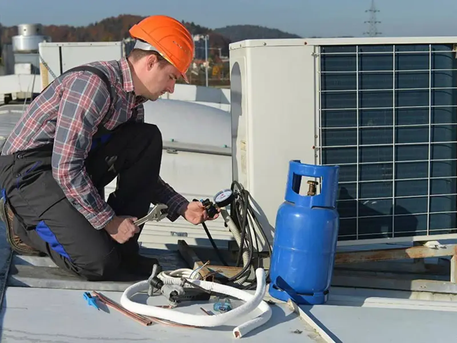 Technician in an orange helmet repairing an air conditioning unit on a rooftop with tools and a blue gas cylinder.