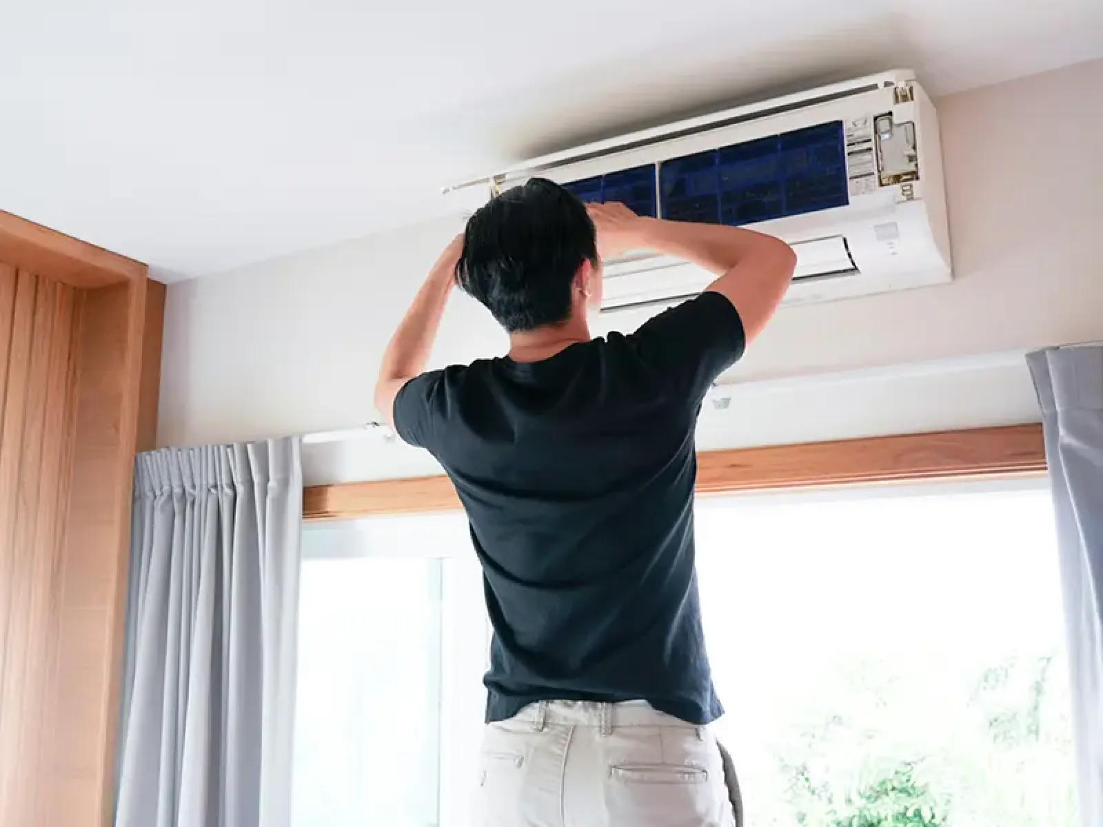 Man in black shirt servicing a wall-mounted air conditioner inside a bright room with curtains and windows.
