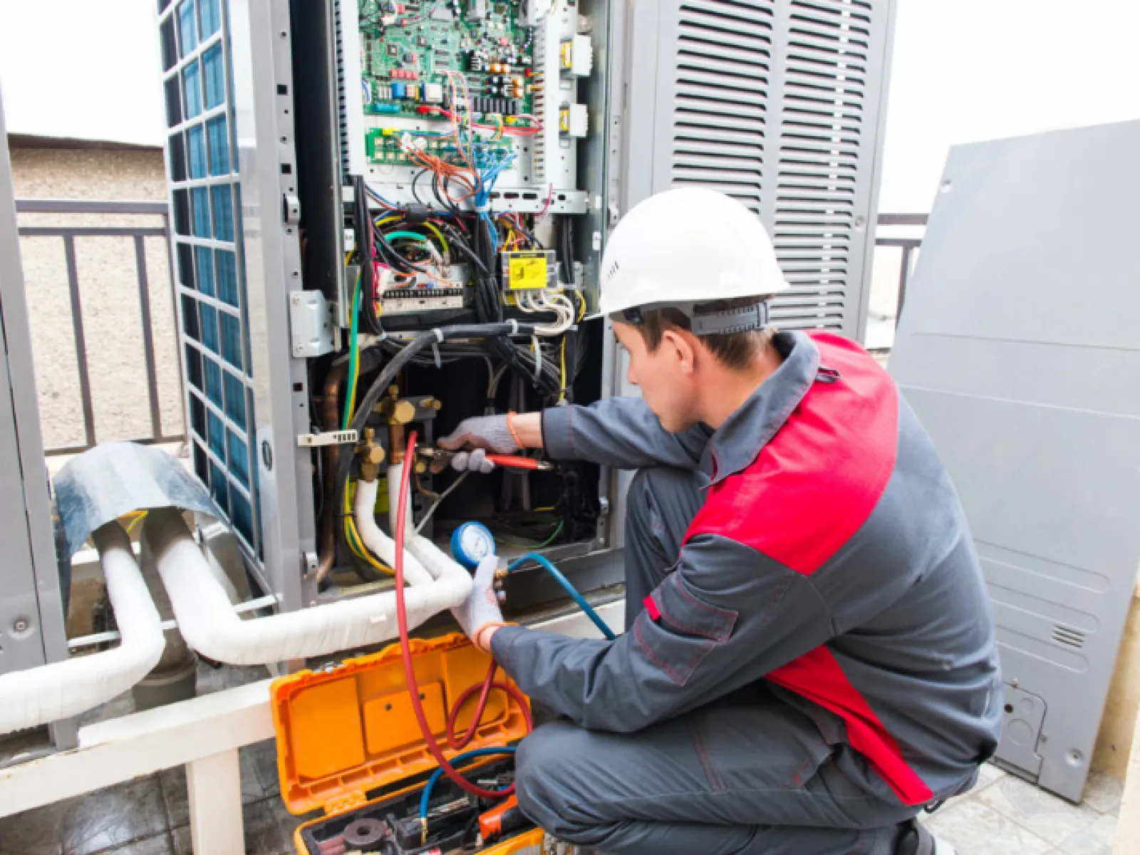 Technician in red and grey uniform repairing and inspecting an outdoor HVAC unit with gauges and tools